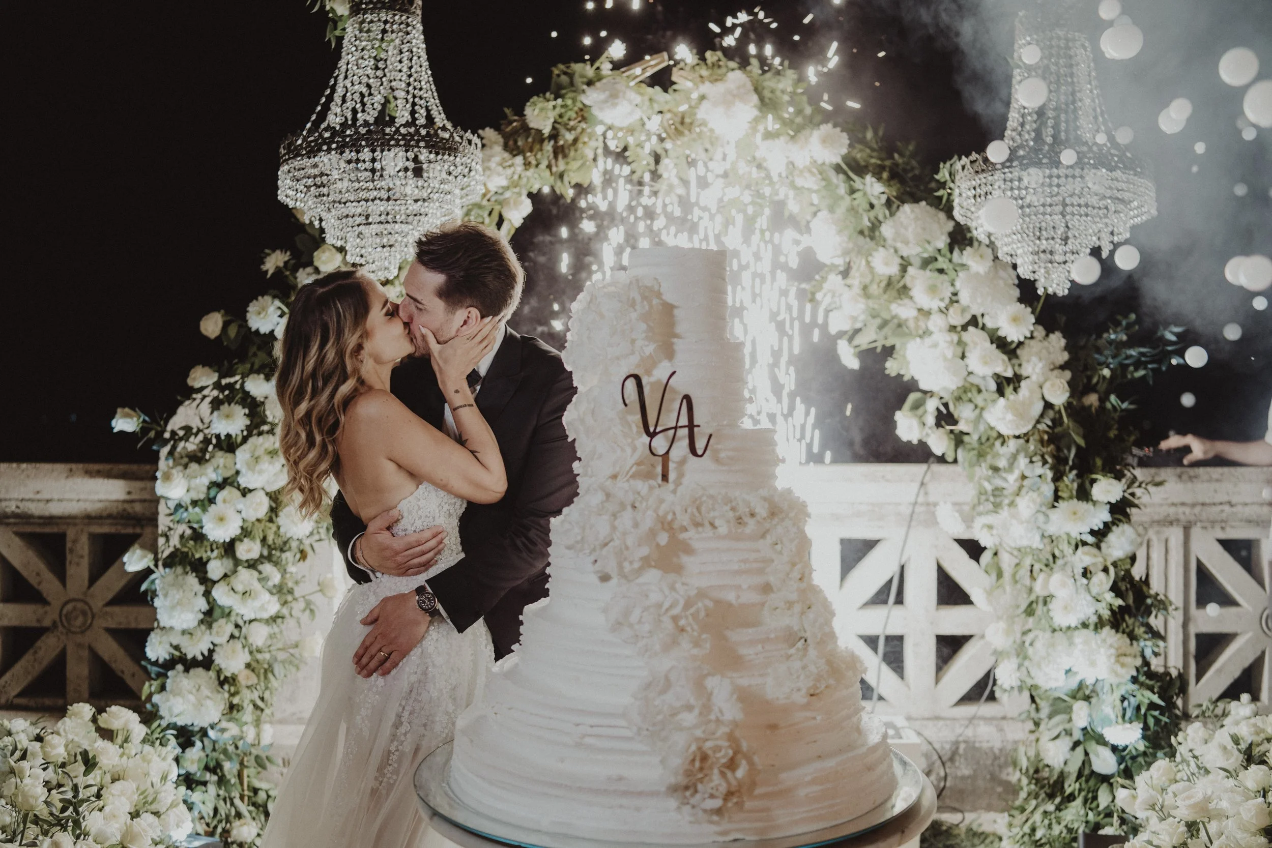Una coppia di sposi si bacia durante il loro matrimonio, circondati da un arco di fiori bianchi e candelabri, con una torta nuziale decorata e accesa alle spalle.