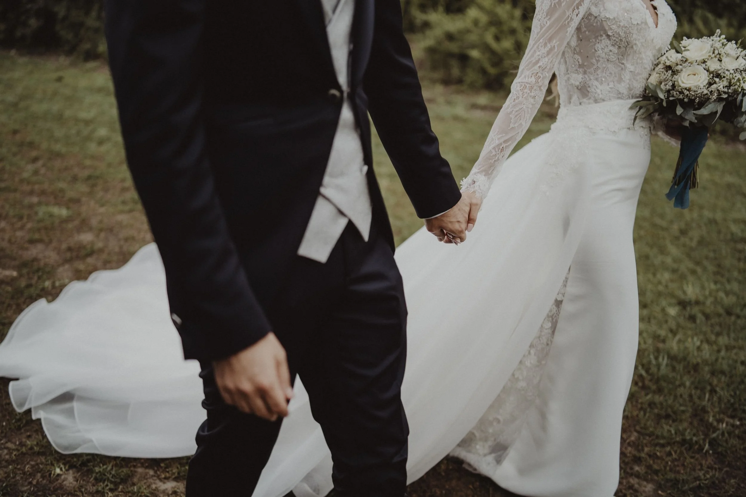Coppia di sposi che si tiene per mano durante il matrimonio all'aperto, con abiti eleganti, sullo sfondo di natura