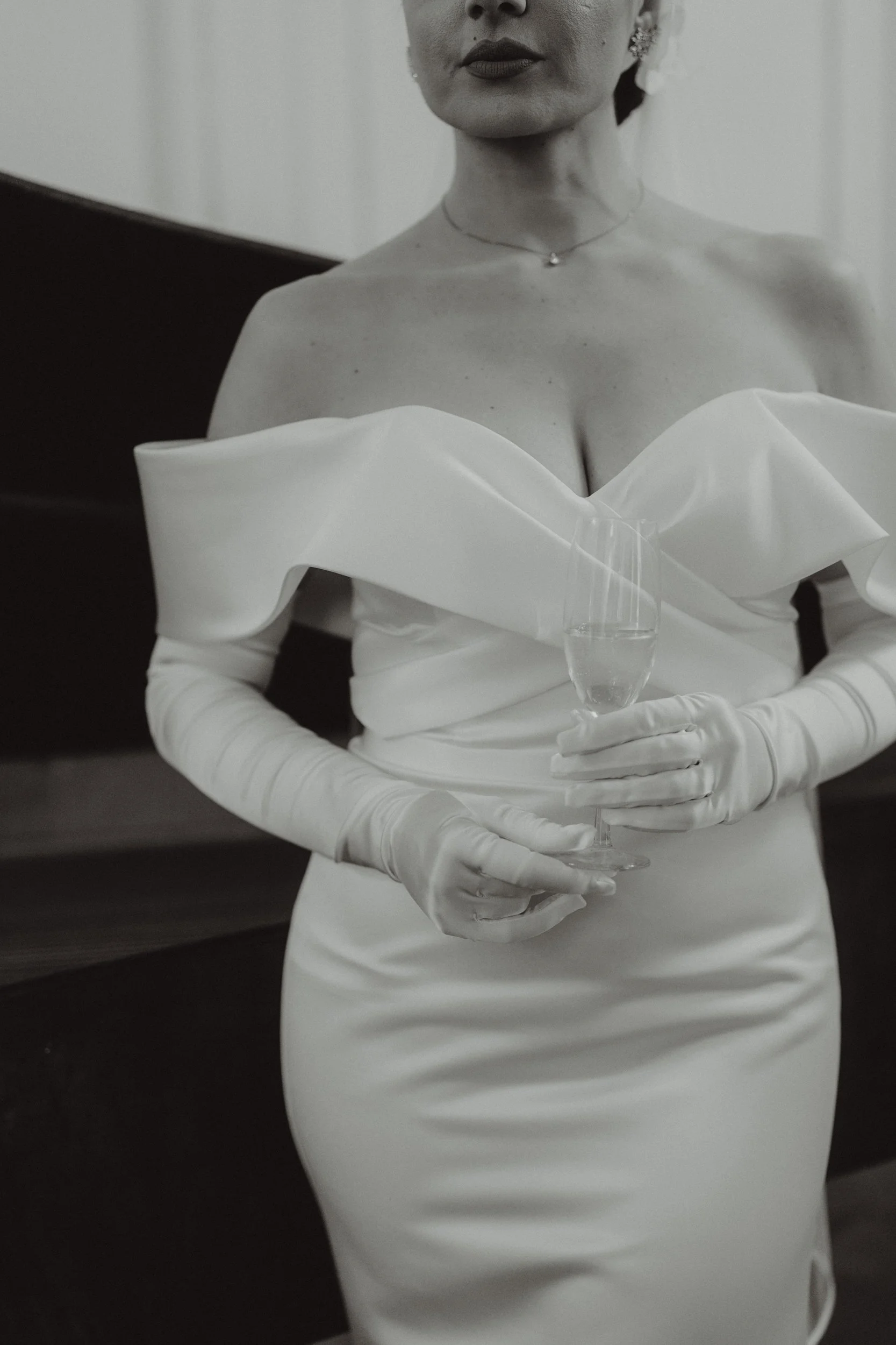Donna in abito da sera bianco, con guanti lunghi, che tiene un bicchiere di champagne mentre posa in un ambiente elegante.