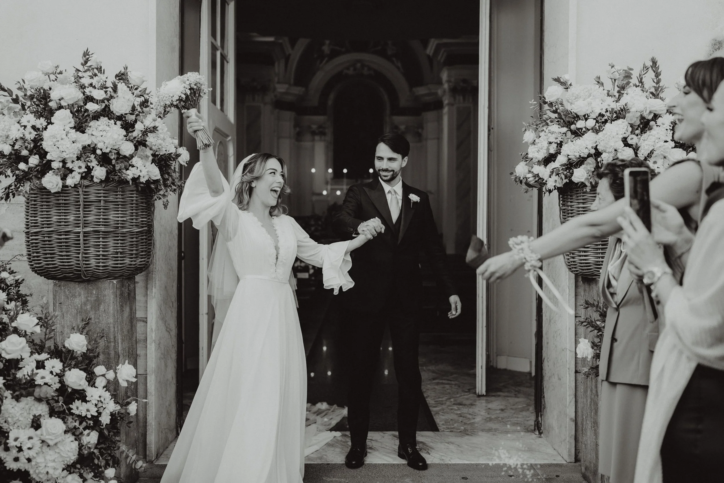 Una coppia di sposi al matrimonio, lei con un vestito bianco e lui in abito nero, escono dalla chiesa tra le coppie di amici che scattano foto e celebrando un momento di gioia.
