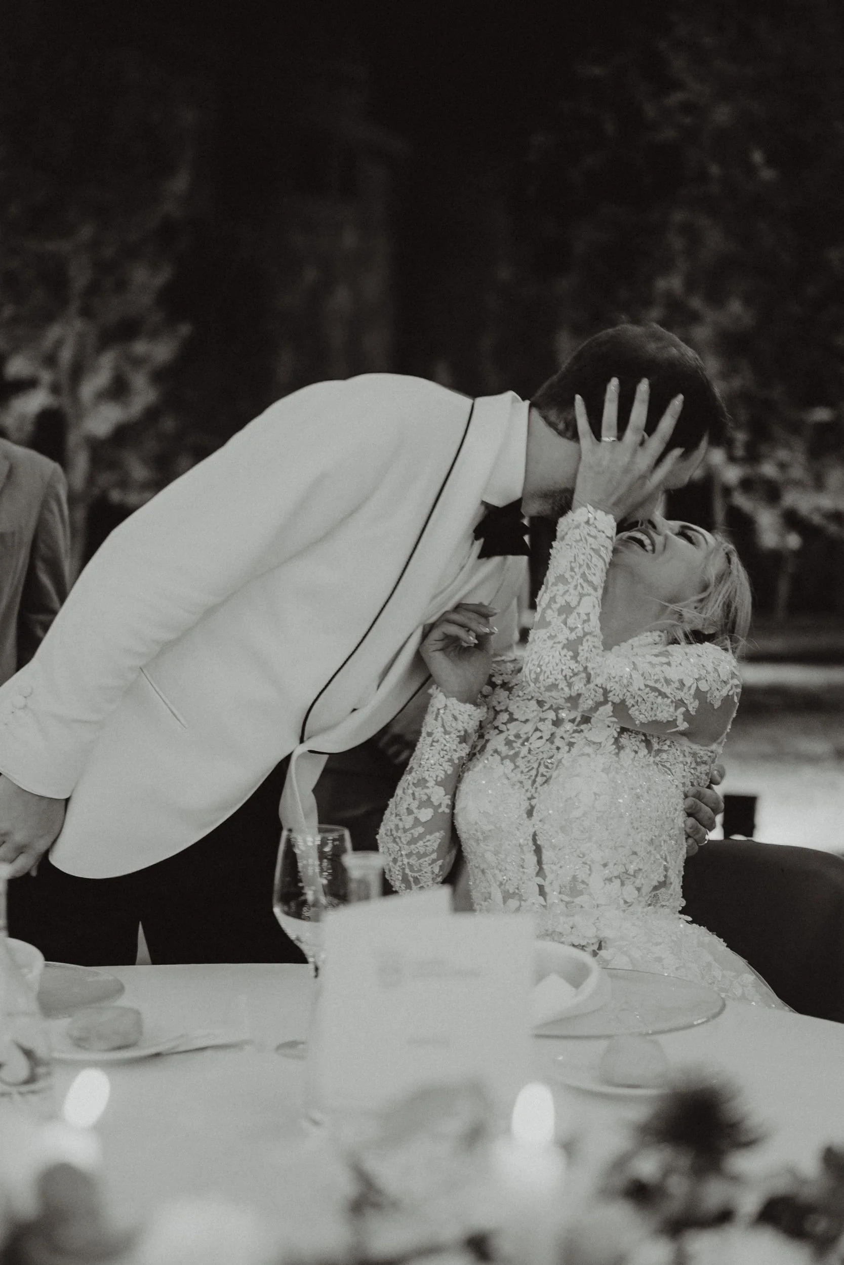 Una coppia di sposi si bacia durante un matrimonio serale, con la donna che sorride e si aggrappa all'uomo in un abito bianco con dettagli in pizzo, mentre lui indossa un completo bianco con papillon.