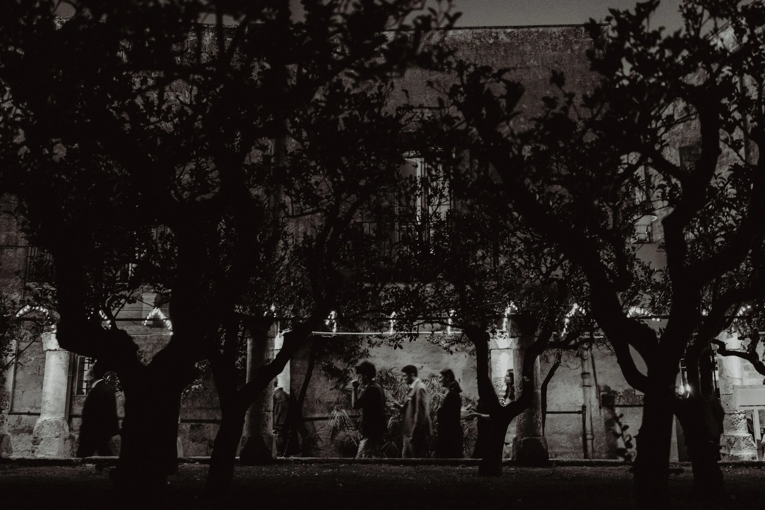 Foto in bianco e nero di cinque persone che camminano tra degli alberi di fronte a un edificio antico, con colonne e pareti di pietra, in un'illuminazione notturna.
