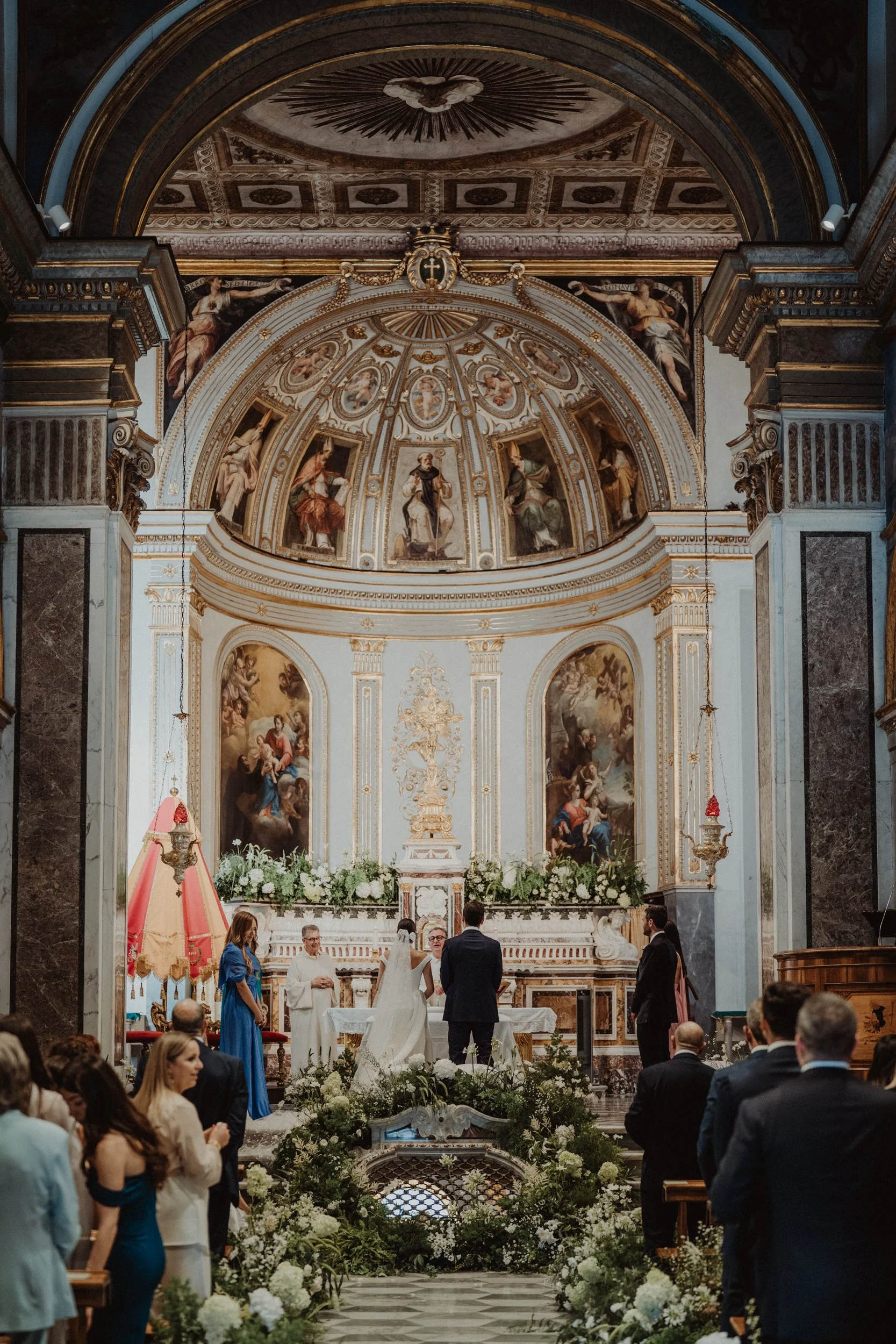 Cerimonia di matrimonio in chiesa con sposi e sacerdote al centro, assistenti e invitati seduti e in piedi, decorazioni floreali bianche e paggetti, in un ambiente barocco con affreschi e arredi dorati.