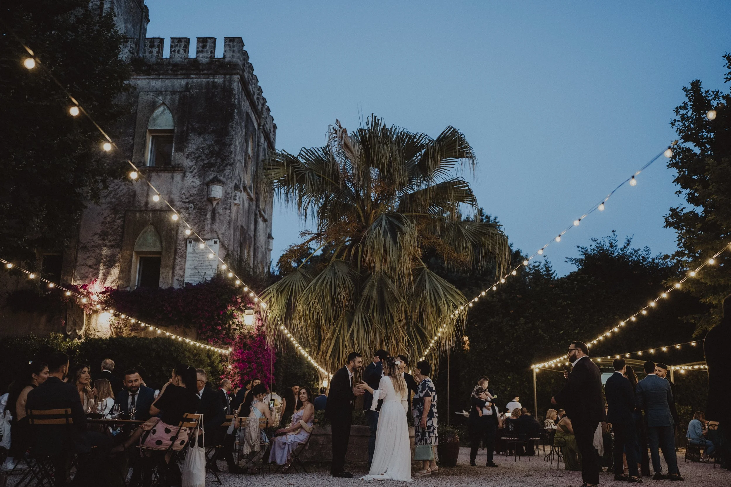 Festa all'aperto di sera con persone in abito elegante, luci decorative e un grande albero al centro, vicino a una vecchia struttura in pietra.