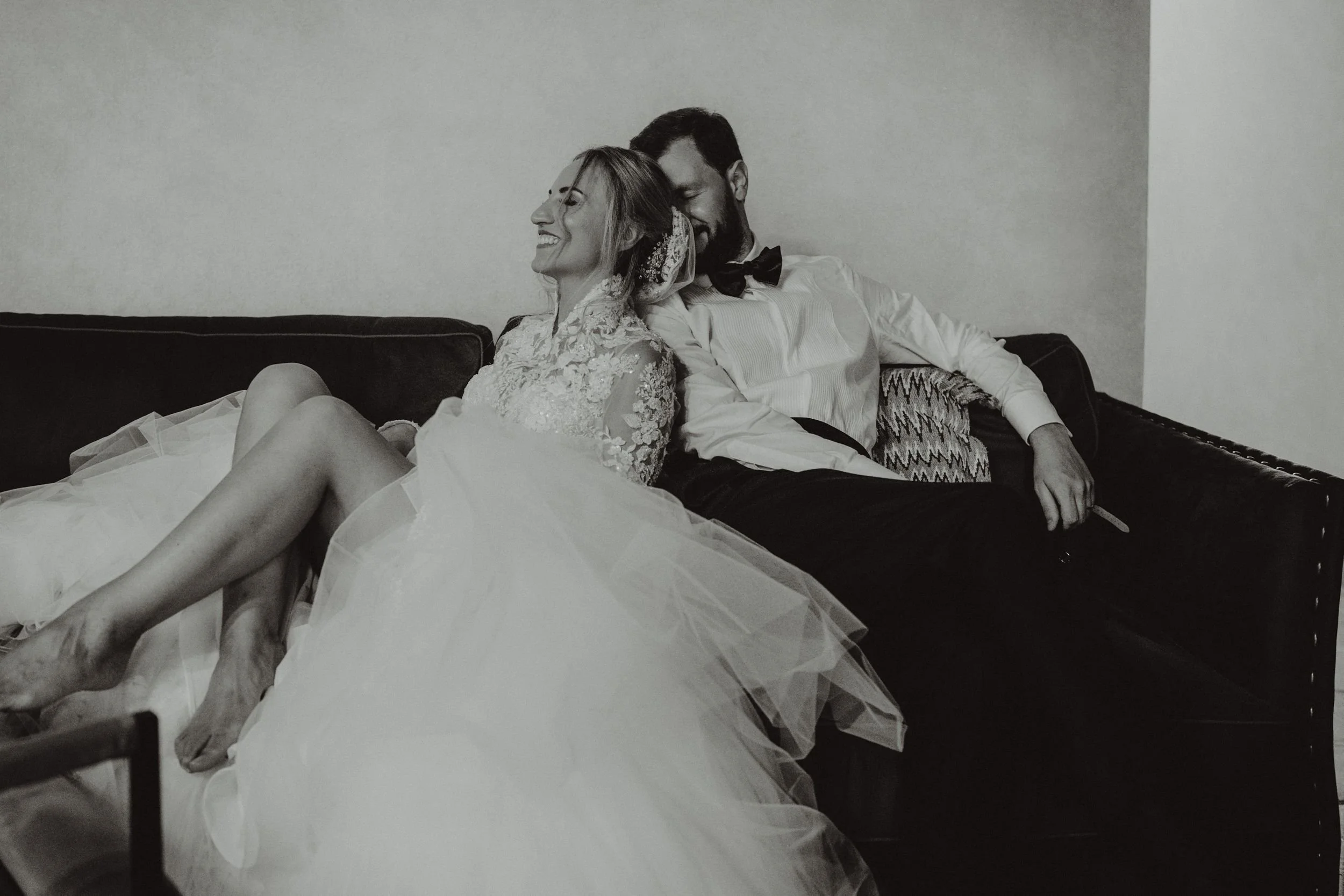 Una coppia di sposi seduti felici sul divano, lei in abito da sposa e lui in abito con papillon, che ridono e si abbracciano in modo affettuoso.