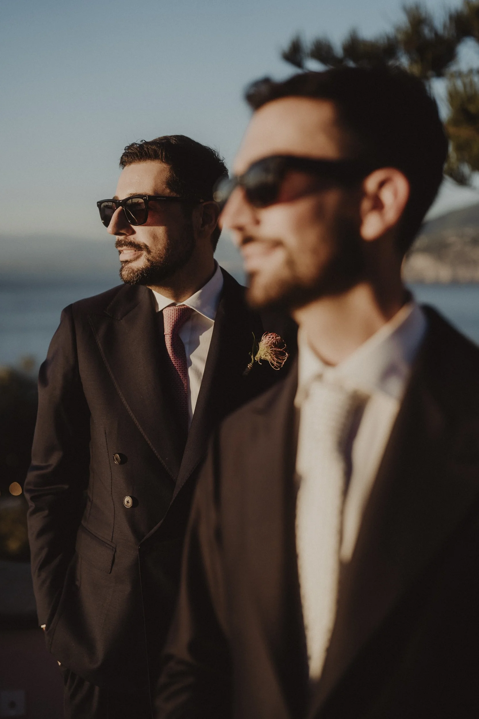 Due uomini vestiti elegantemente con giacche nere, occhiali da sole e cravatte, uno più in primo piano e l'altro più lontano, che si godono un tramonto sulla costa con il mare e le colline sullo sfondo.