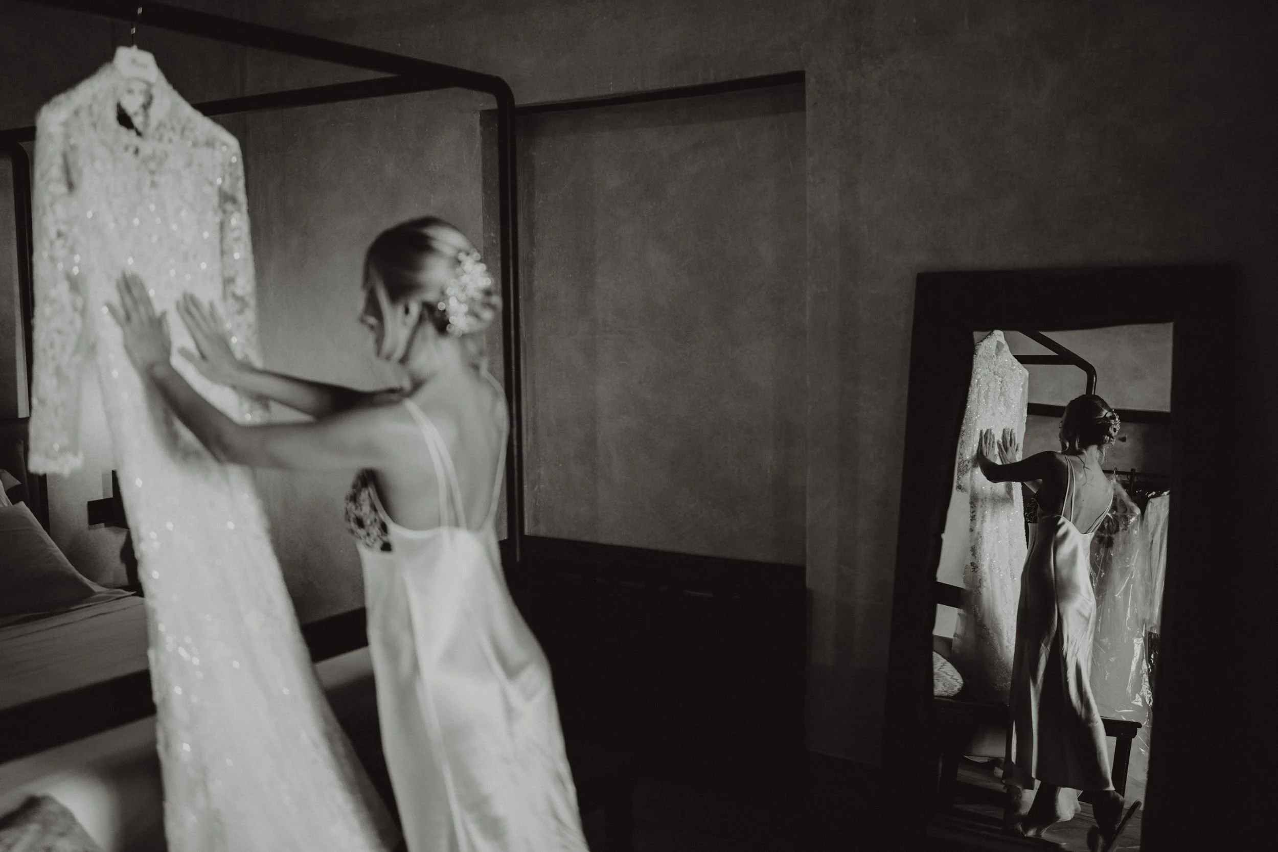 Una donna con un vestito da sposa guarda un abito da sposa appeso, mentre si specchia in uno specchio grande in una stanza.