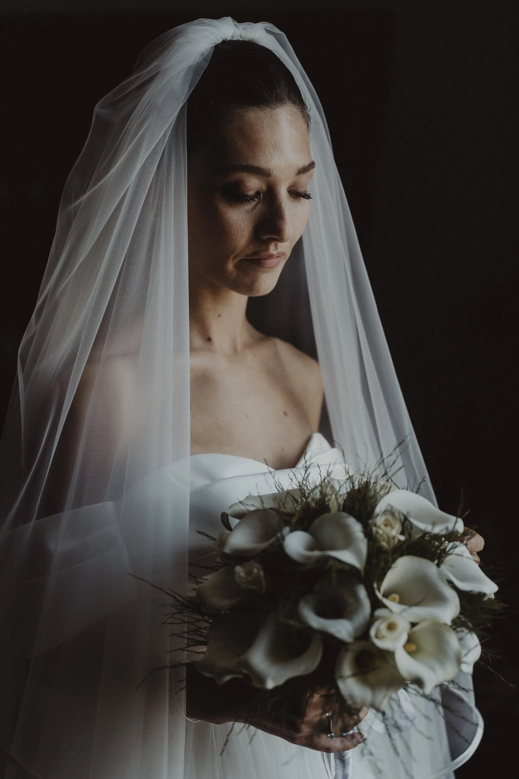 Una sposa con velo bianco e abito bianco, che guarda un bouquet di fiori bianchi, su uno sfondo scuro.