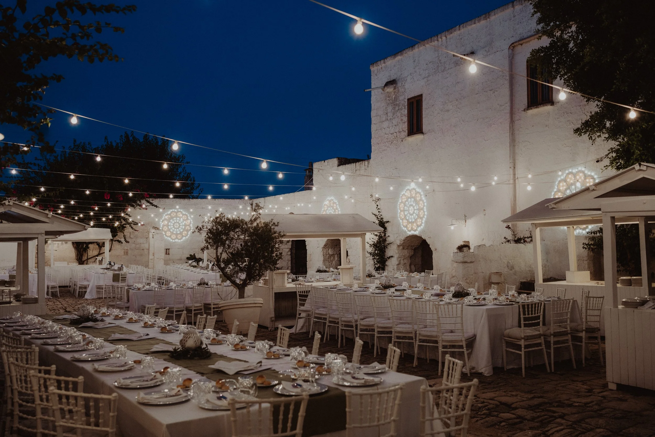 Cena all'aperto con tavoli imbanditi, luci decorative e decorazioni luminose su un muro bianco, in un'atmosfera serale.