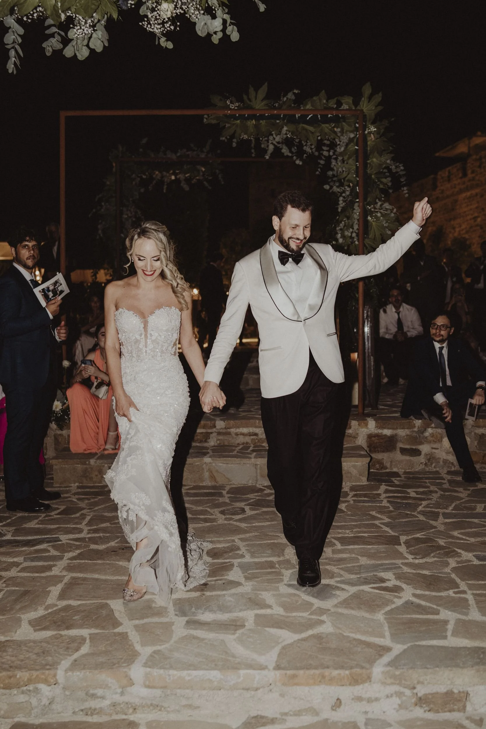 Una coppia di sposi danza sorridendo durante un matrimonio notturno, circondati da invitati.