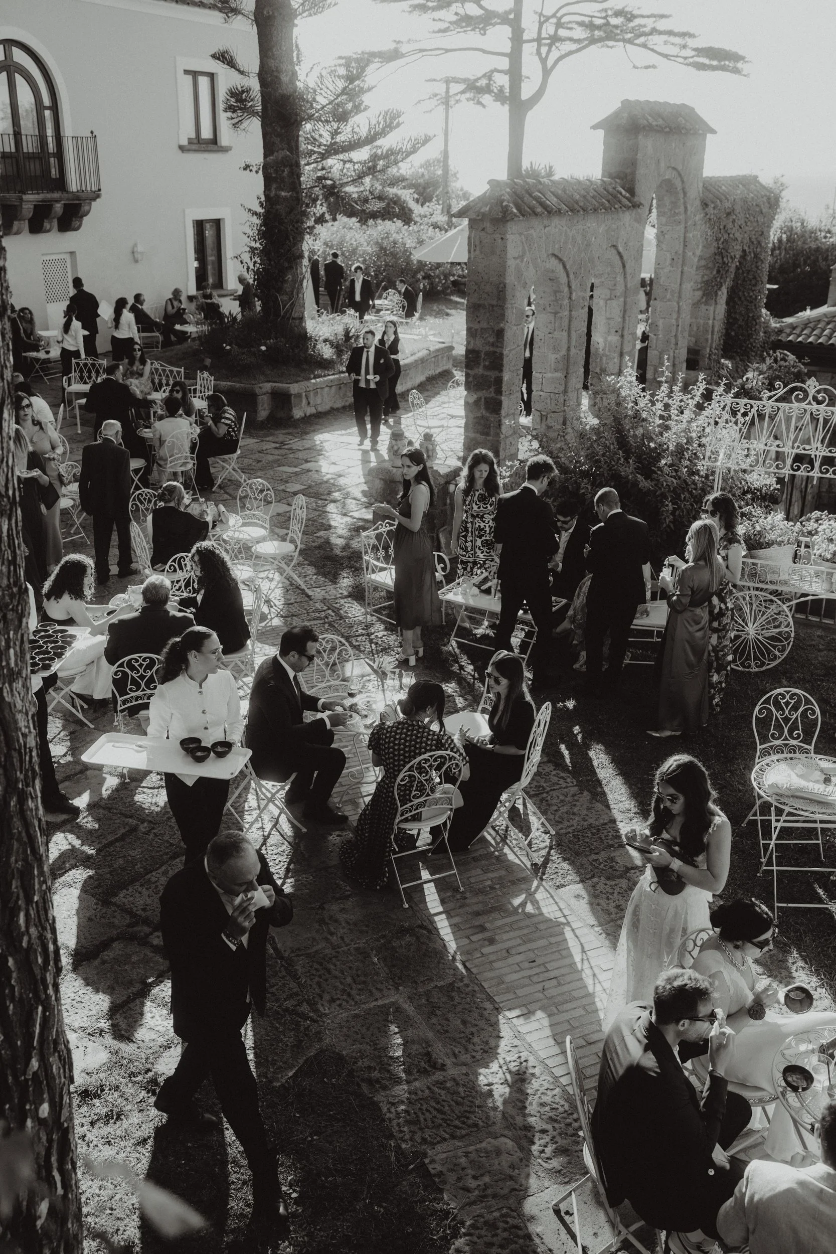 Una scena di ritrovo all'aperto con molte persone eleganti sedute e in piedi, che si godono il sole e una festa all'aperto in un cortile con archi in pietra e alberi, in bianco e nero.