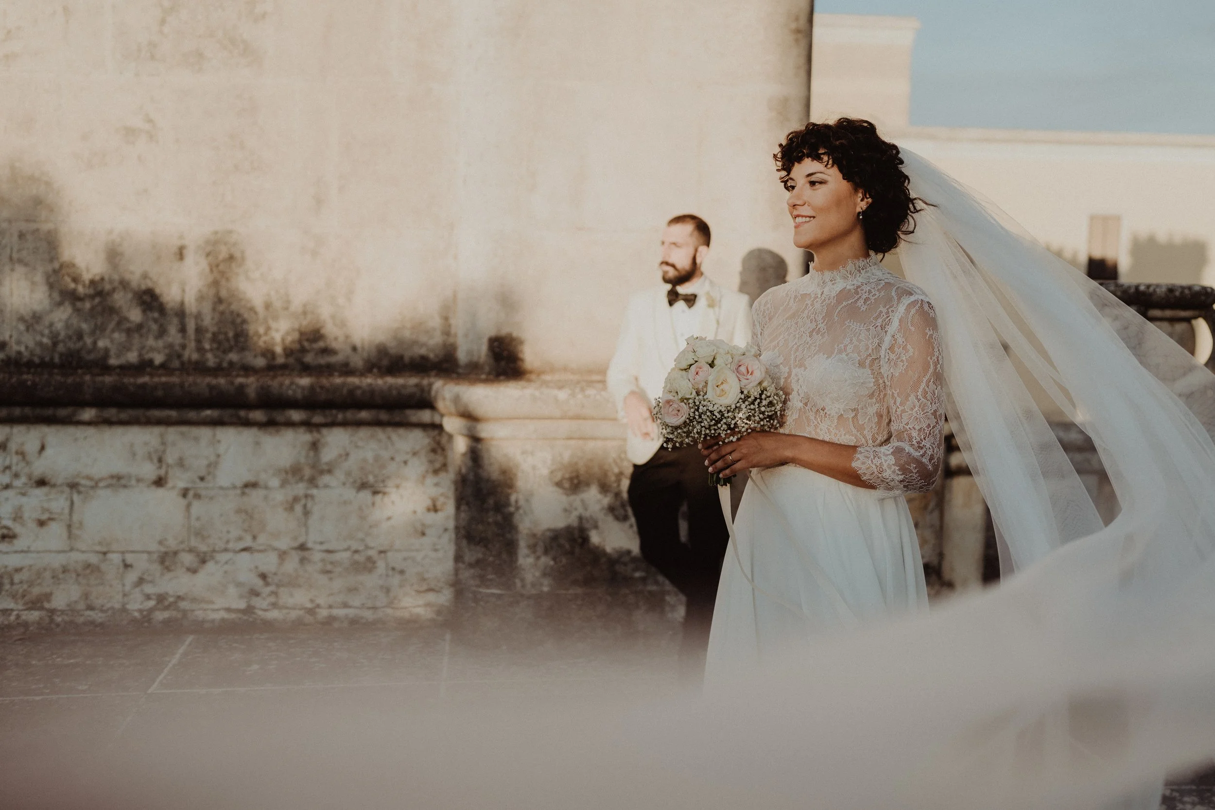 Una sposa in abito da sposa con velo, che sorride e tiene un bouquet di fiori, in un luogo all'aperto con un muro di pietra sullo sfondo, mentre un uomo in abito bianco e pantaloni neri si trova dietro di lei.