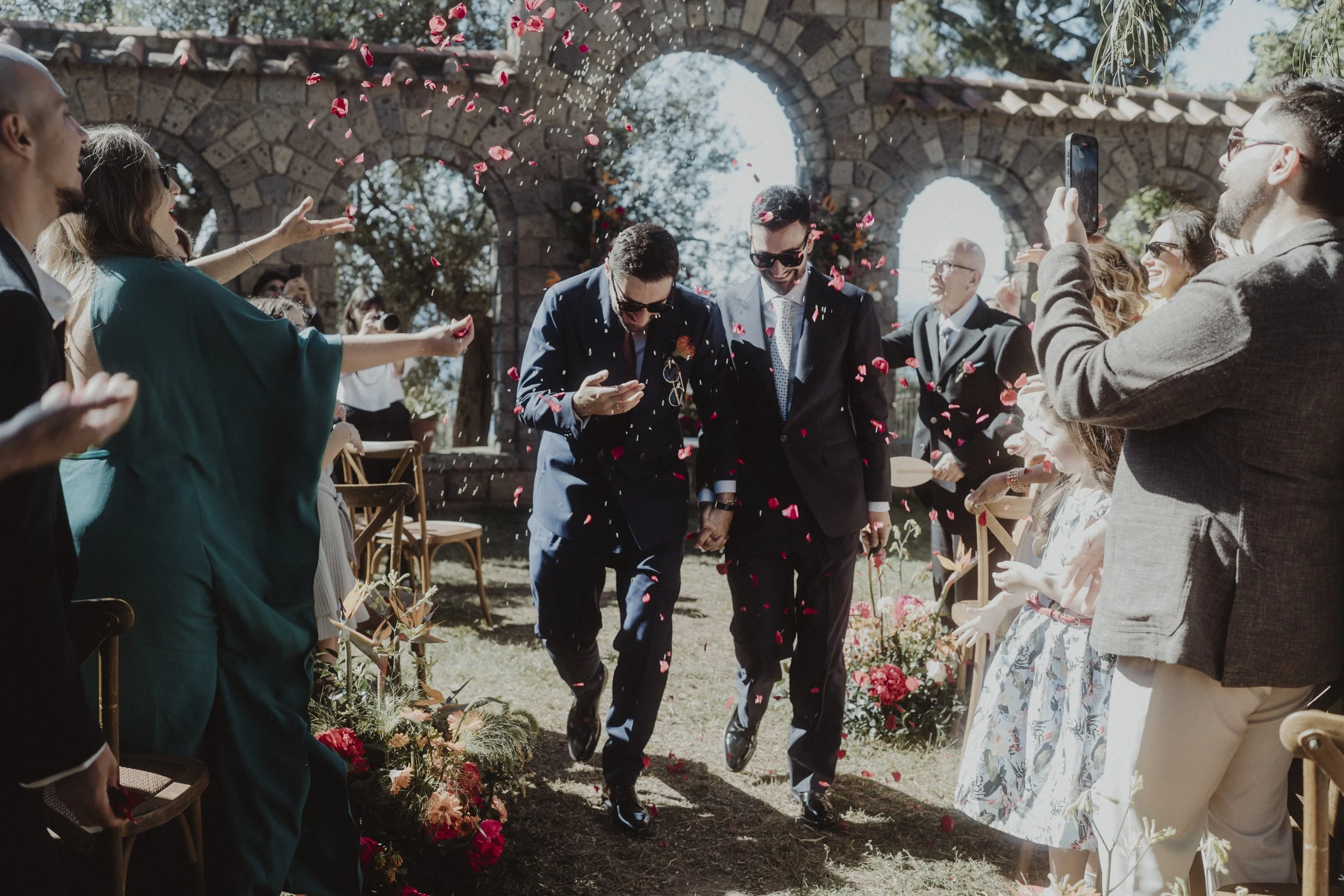Due uomini in abiti da matrimonio camminano mano nella mano tra amici e parenti che li festeggiano con petali di rosa in un cortile con archi in pietra, durante il matrimonio.