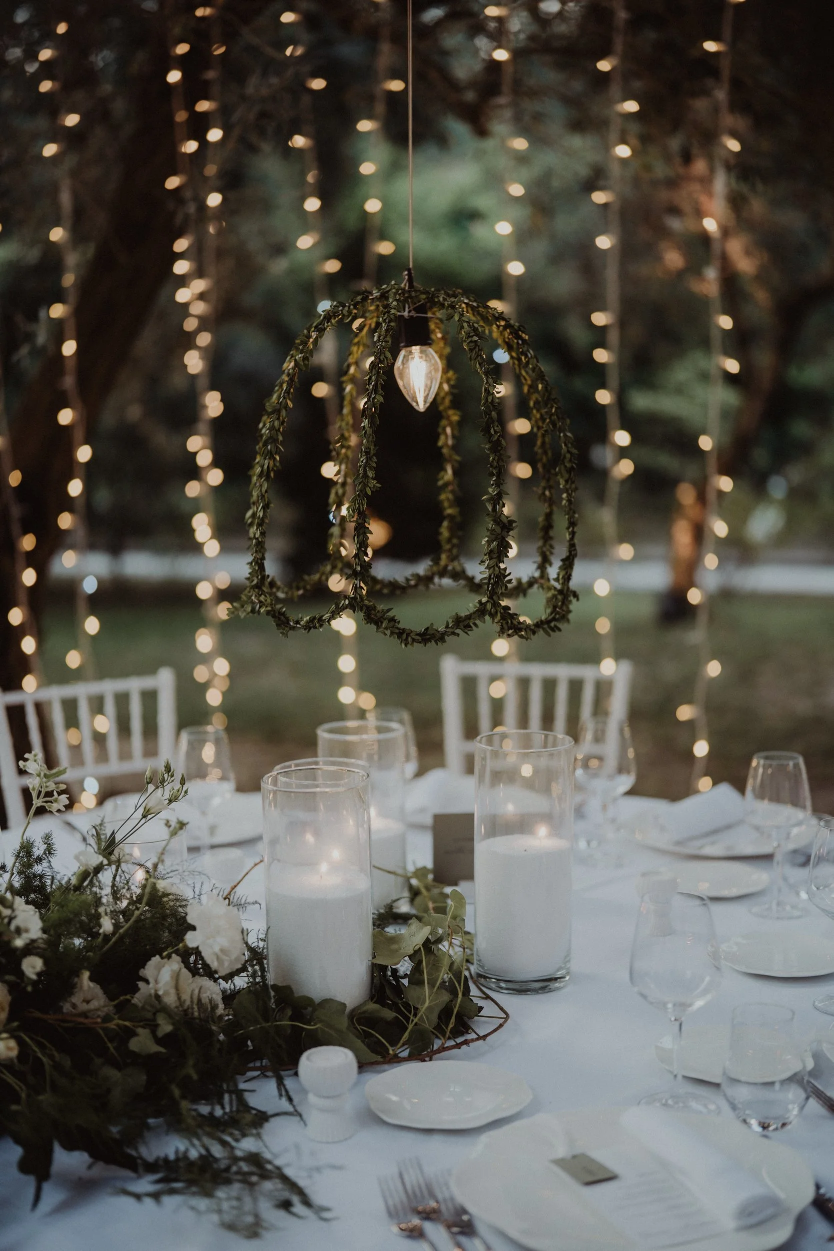 Tavolo imbandito con candele, fiori e decorazioni, sotto luci di stringa, in un ambiente esterno.