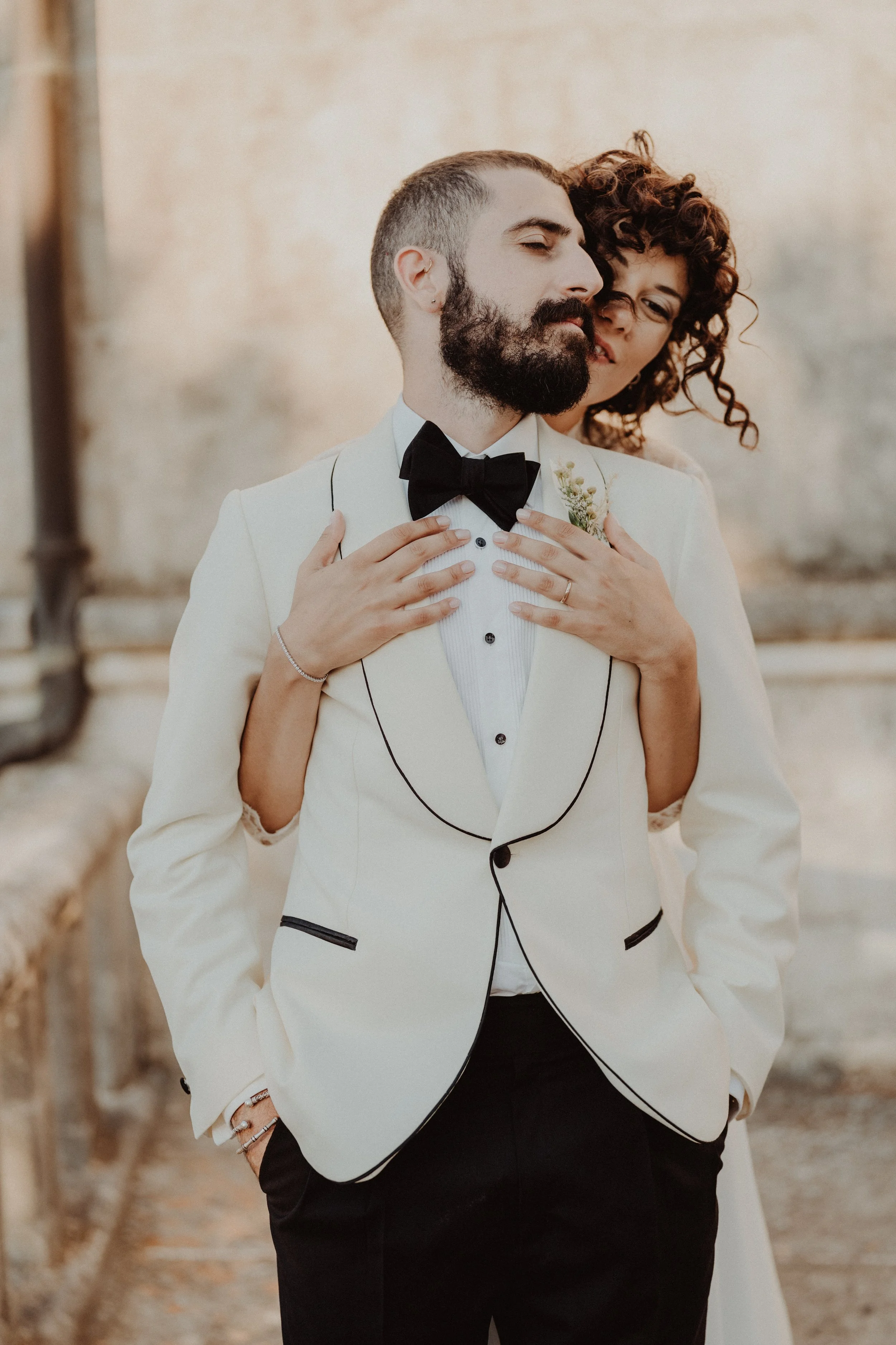 Una coppia di sposi in abiti eleganti, l'uomo indossa un completo bianco con collo a revers nero e una cravatta nera, mentre la donna si appoggia alle sue spalle e lo abbraccia. Sono in piedi in un ambiente rustico con pareti di pietra.