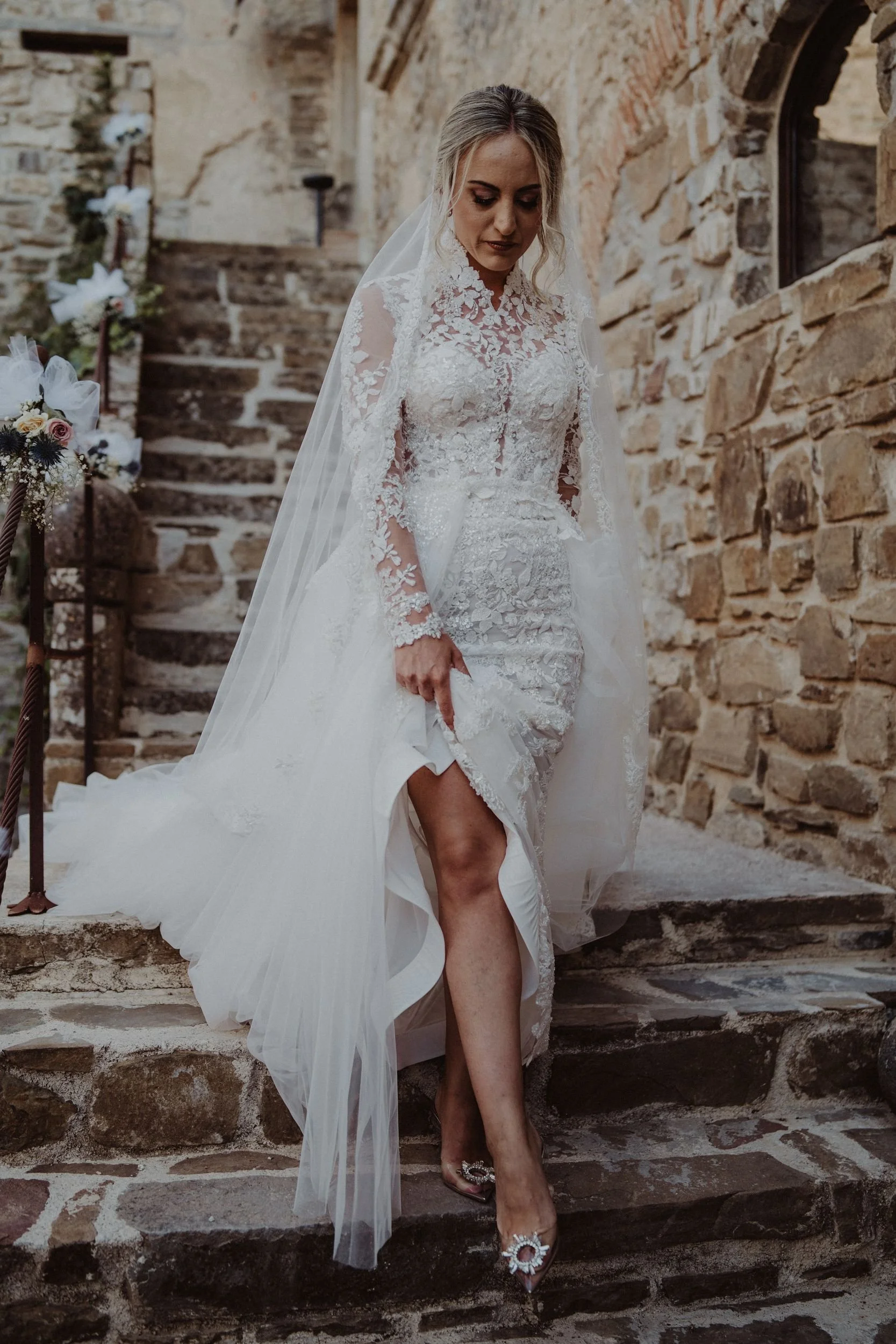 Una sposa in abito da matrimonio bianco con dettagli in pizzo, che scende le scale di pietra in un ambiente storicamente in pietra, con decorazioni floreali.