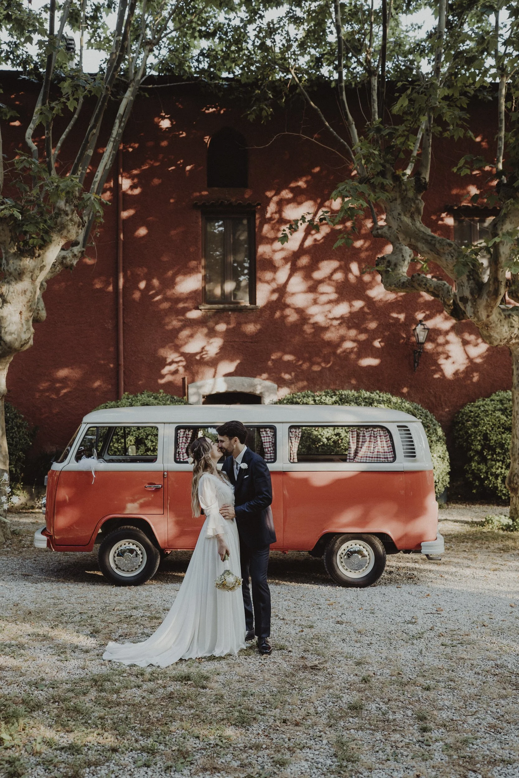 Una coppia di sposi in abiti da matrimonio si bacia davanti a un furgone vintage di colore rosso e bianco, con alberi e un edificio rosso sullo sfondo.