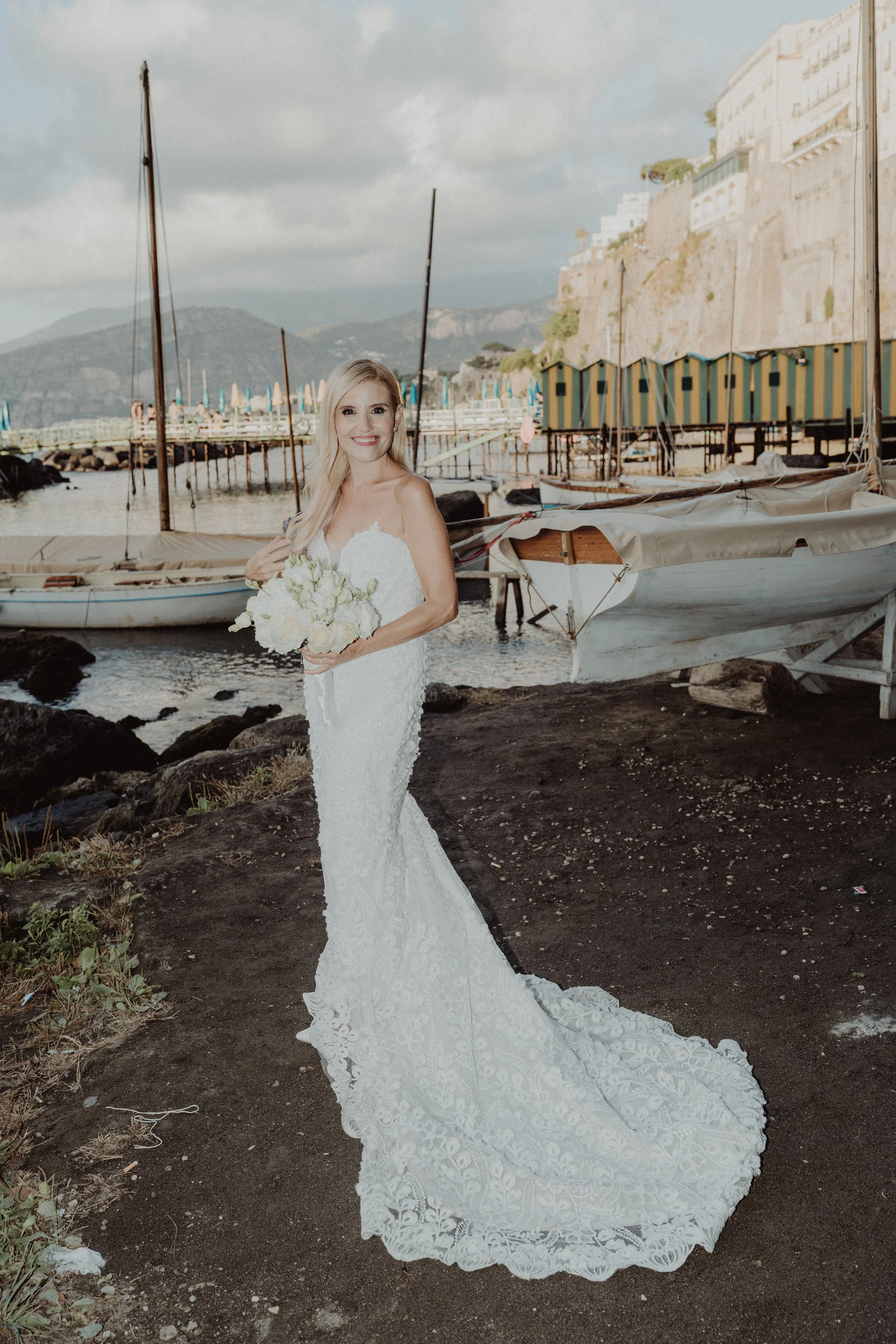 Una sposa in abito da sposa bianco con bouquet di fiori bianchi, si trova sulla spiaggia vicino a barche e un molo, con un paesaggio di scogli e edifici sullo sfondo.