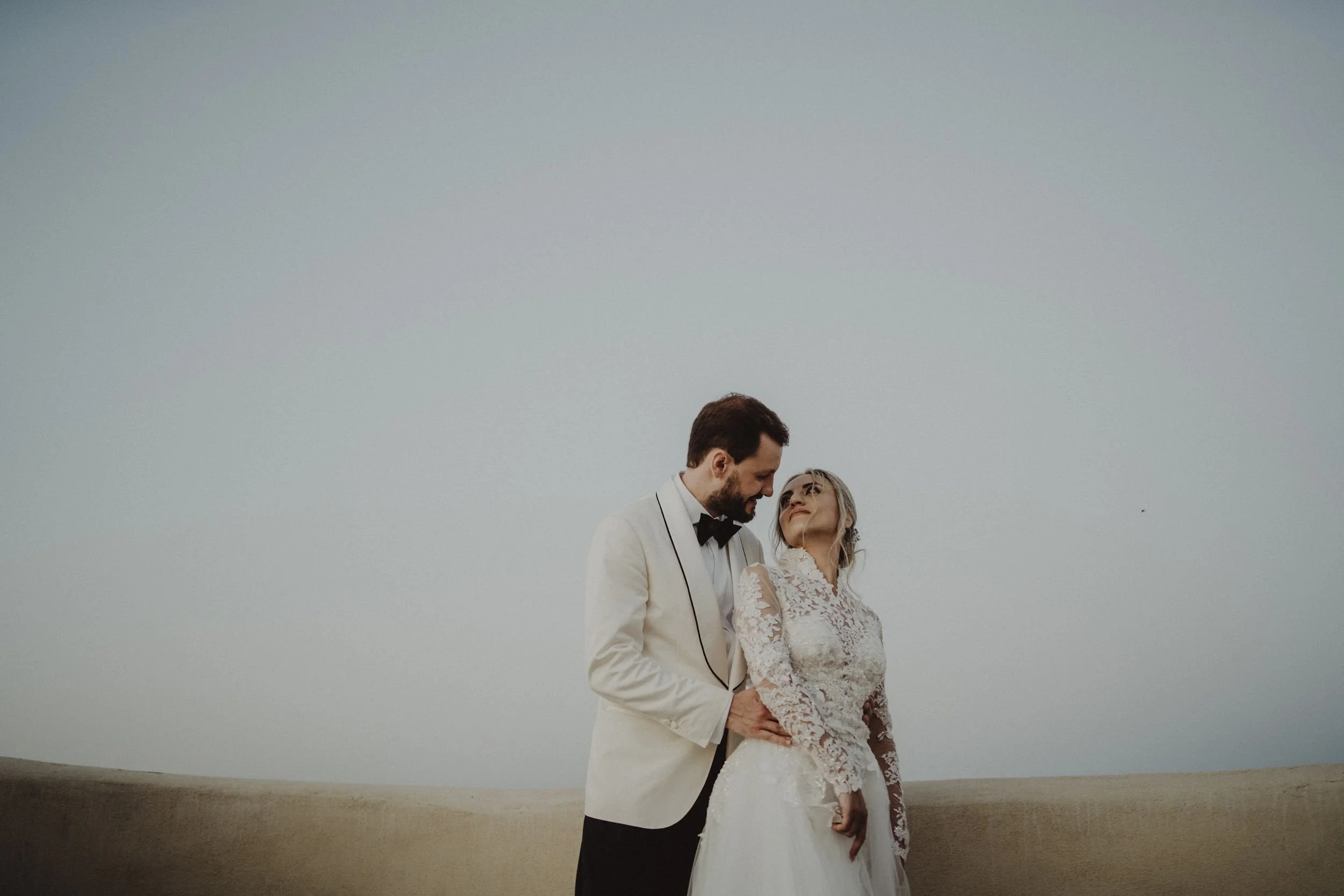 Una coppia di sposi in abito da matrimonio si guarda amorosamente in un paesaggio senza nuvole.