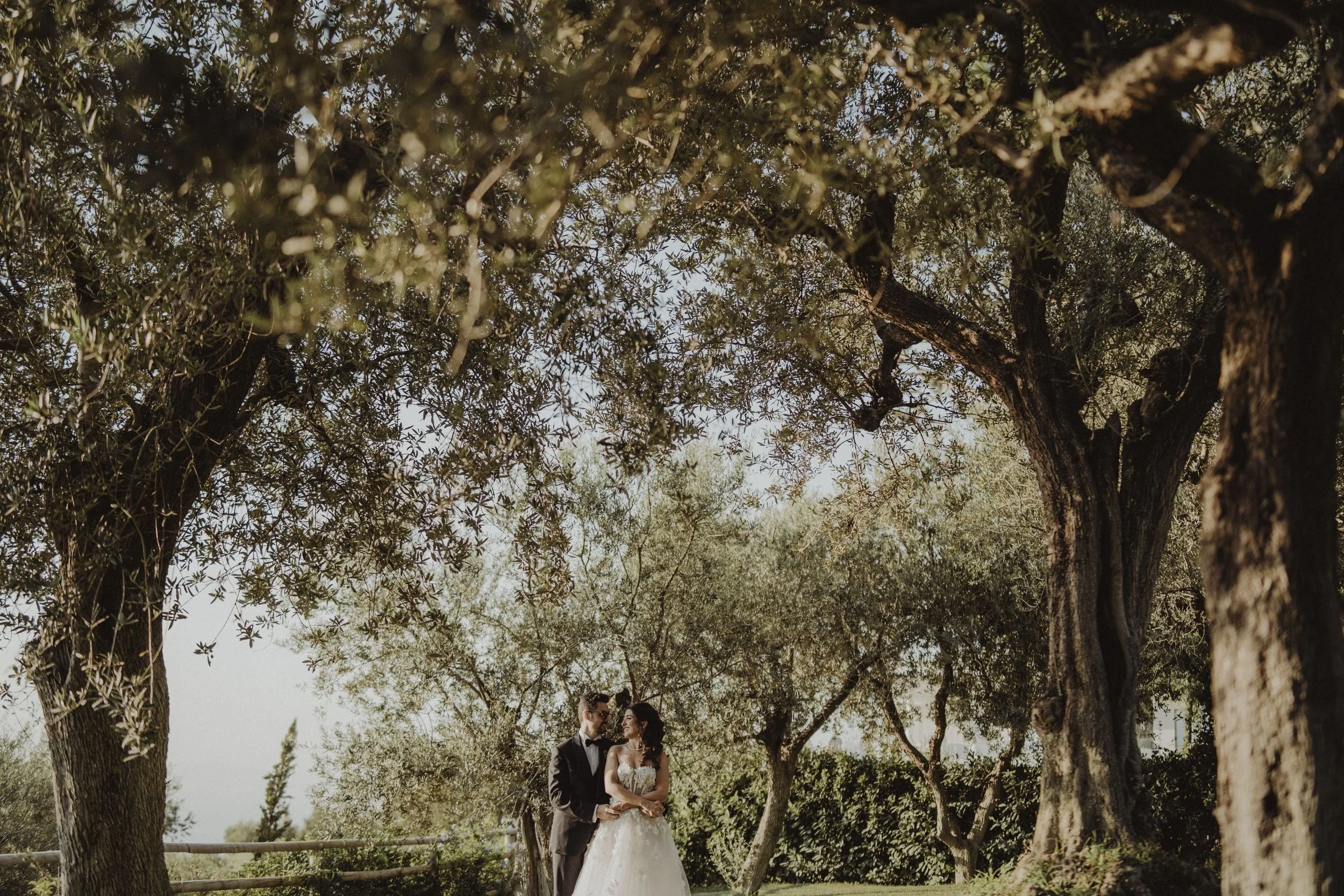 Una coppia di sposi in abiti eleganti in un giardino tra alberi, scattata durante un matrimonio all'aperto al tramonto.