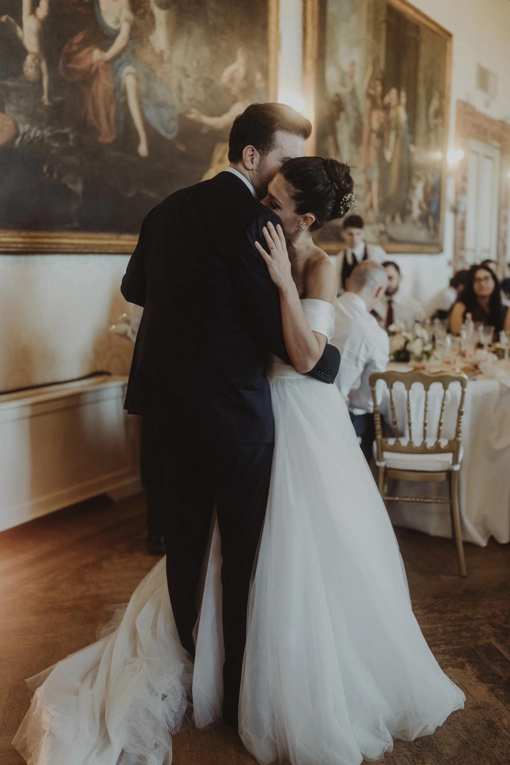 Una coppia di sposi si abbraccia durante il matrimonio in un'elegante sala da banchetto, con un quadro dipinto alle spalle e tavoli decorati con fiori e candele.