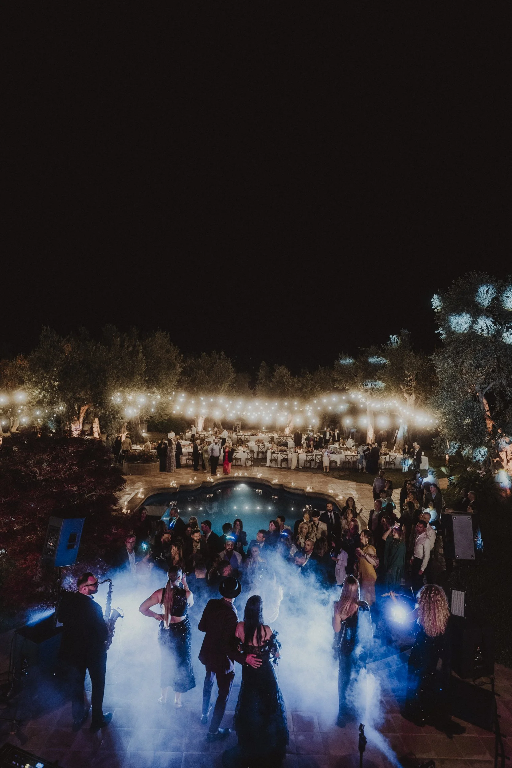 Festa serale all'aperto con persone che ballano su una pista vicino a una piscina, decorazioni luminose e tavoli apparecchiati in un giardino.