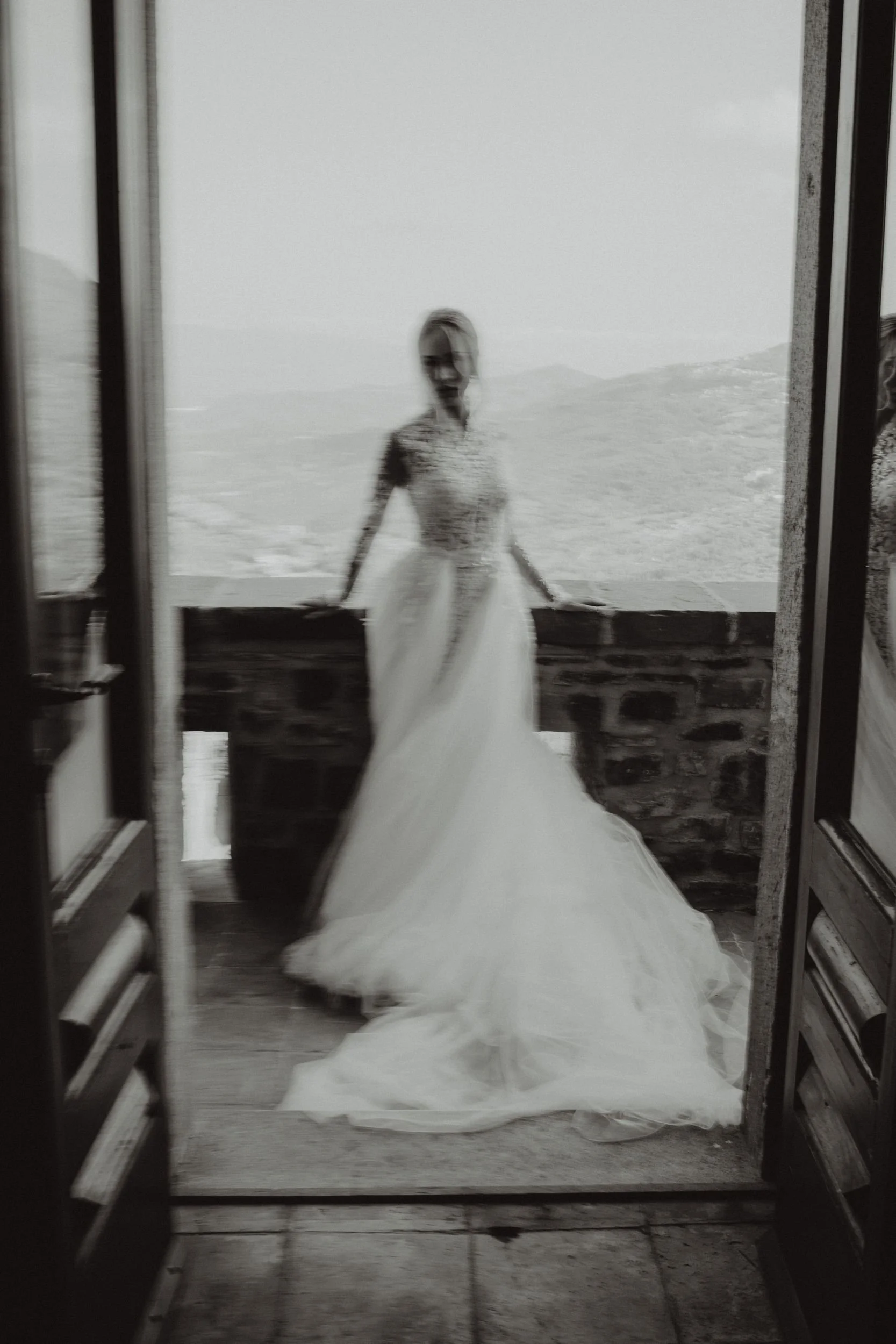 Donna in abito da sposa che si trova su un balcone o terrazza con vista su una valle montuosa in background.