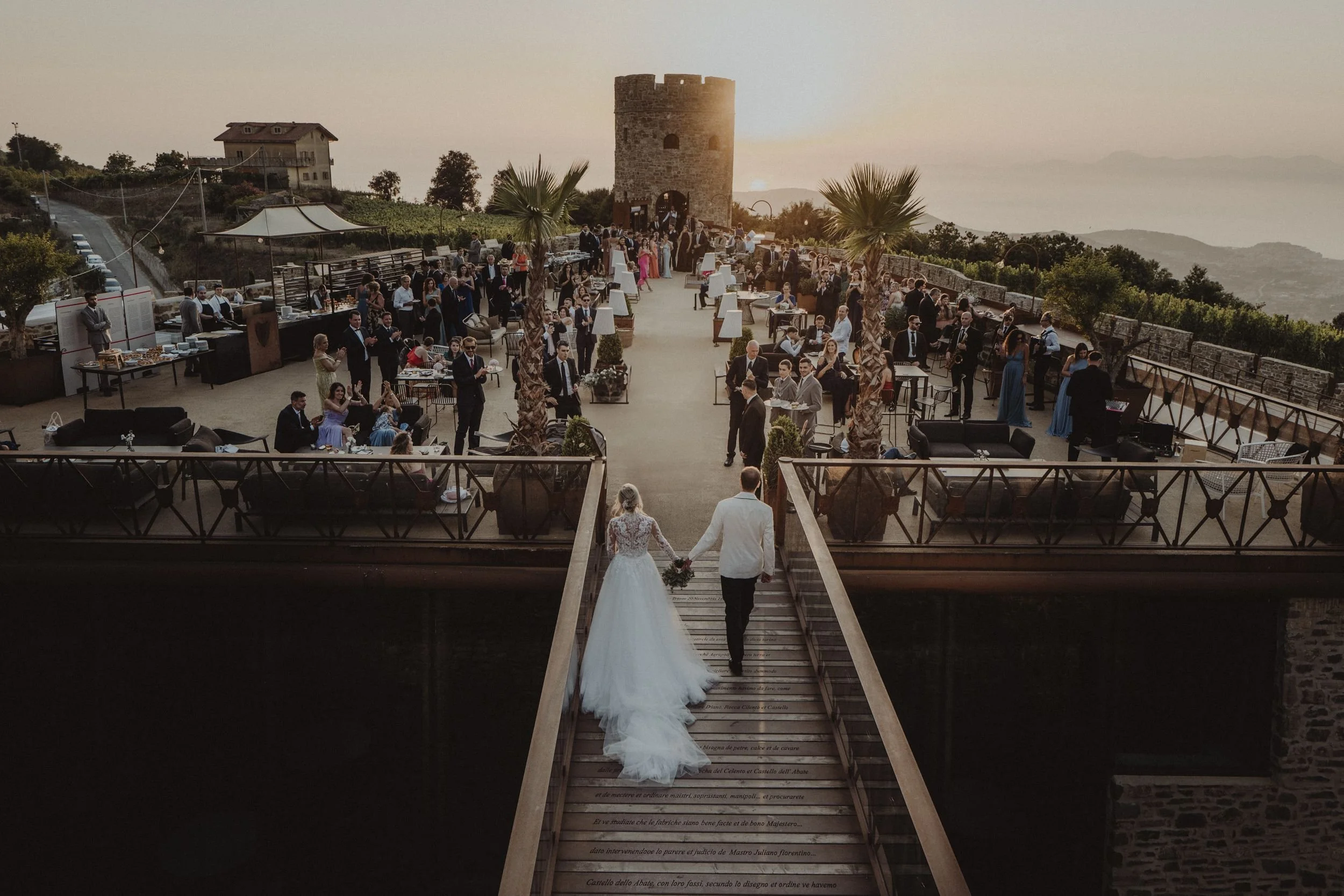 Cerimonia di nozze in un terrazzo con vista al tramonto, con sposi che camminano mano nella mano sul ponte di legno. Ospiti seduti e in piedi, musica dal vivo, un castello e un paesaggio montano sullo sfondo.