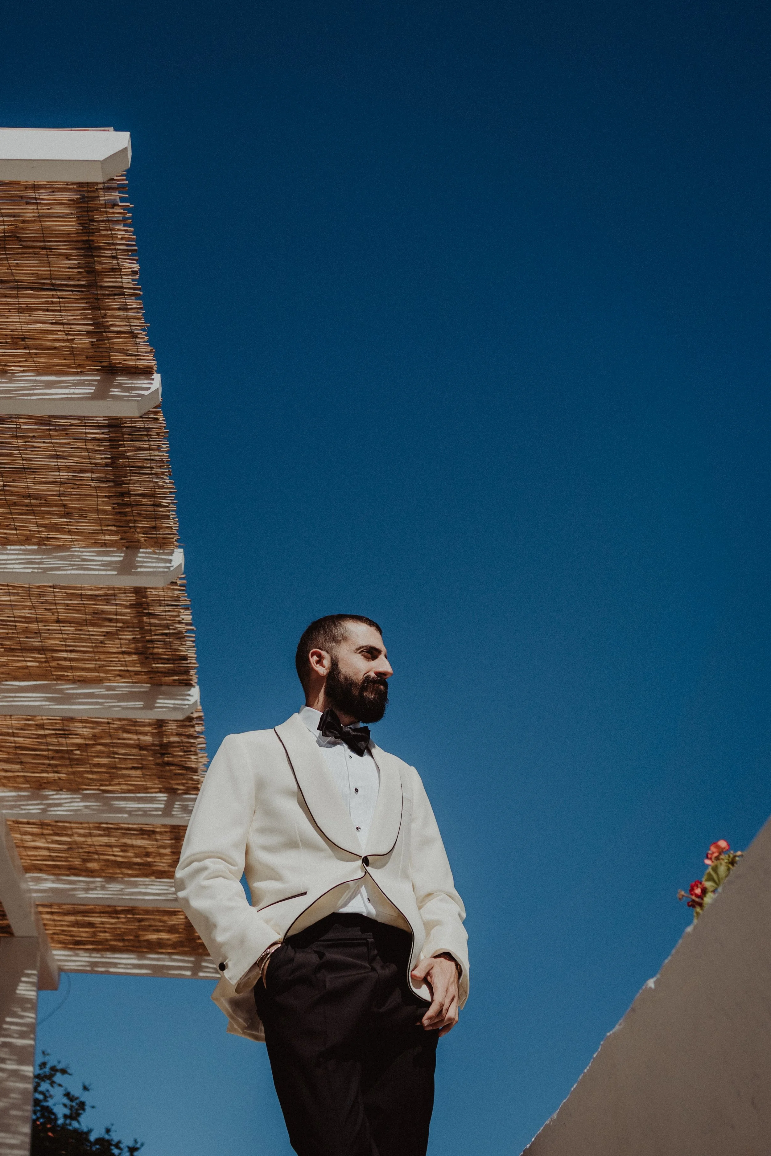 Uomo con abito bianco e smoking nero, con barba, che sorride all'aperto con cielo blu come sfondo.