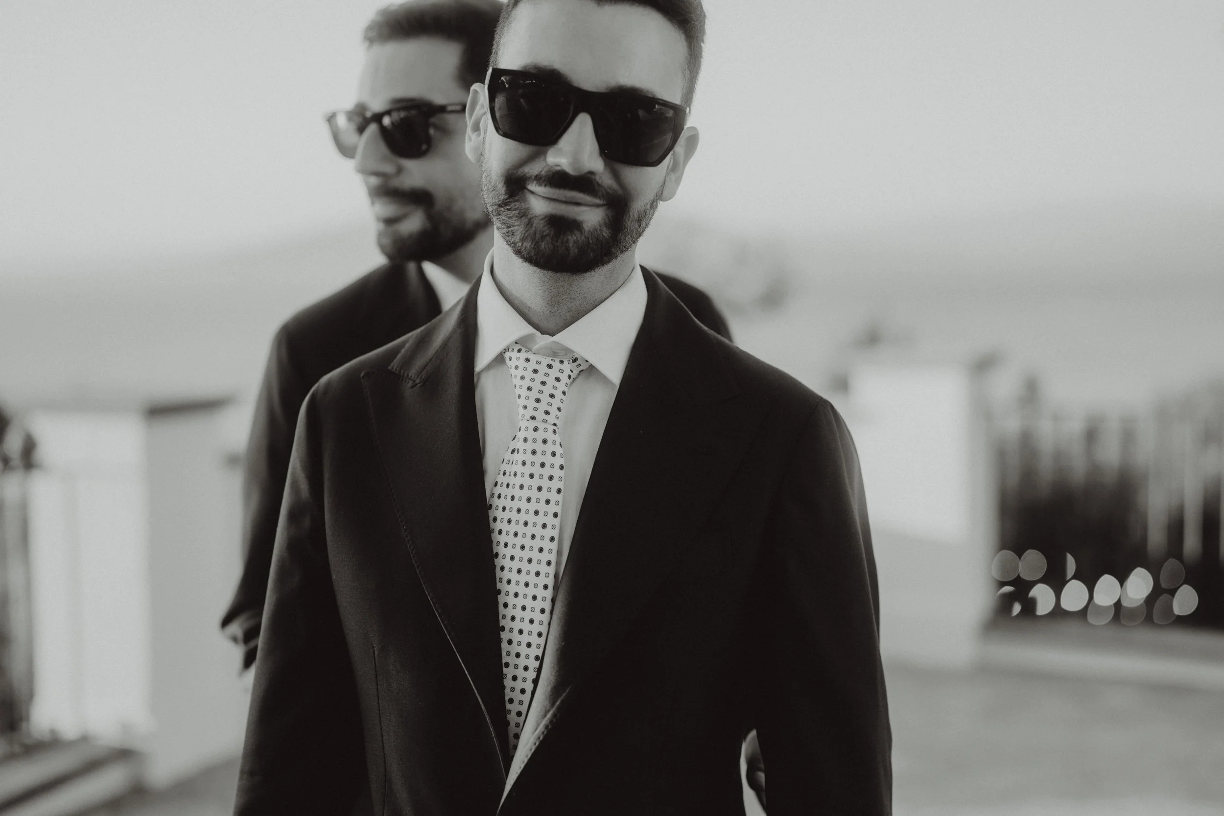 Due uomini vestiti elegantemente con abiti e occhiali da sole che camminano insieme all'aperto, sfondo sfocato.