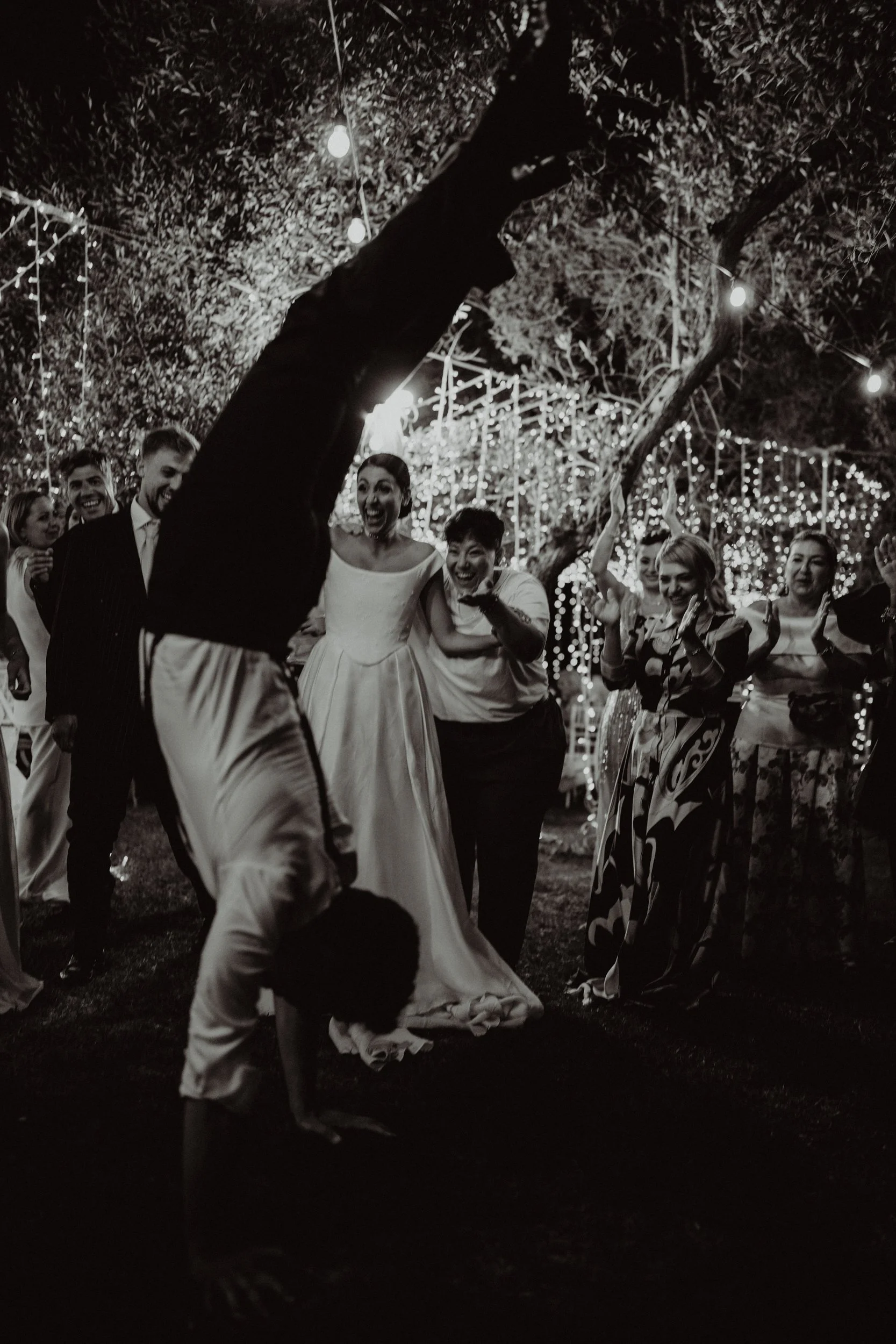 Gruppo di persone che festeggiano un matrimonio, con un uomo che fa il handstand e una sposa sorridente, decorazioni luminose di luci natalizie nel giardino.
