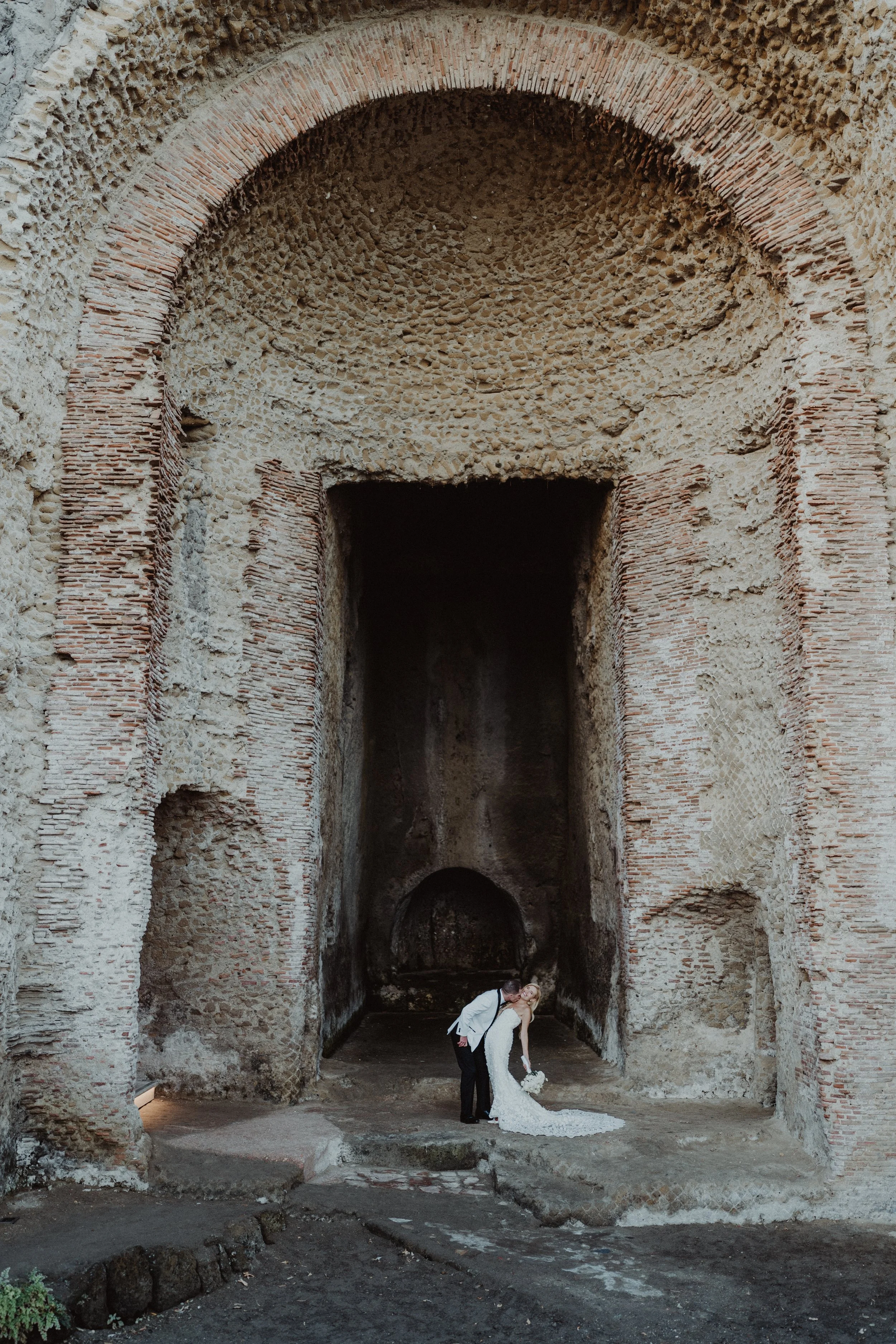 Una coppia di sposi in abiti da matrimonio si bacia davanti a una grande parete di rovine antiche, con archi e muri di pietra e mattoni.