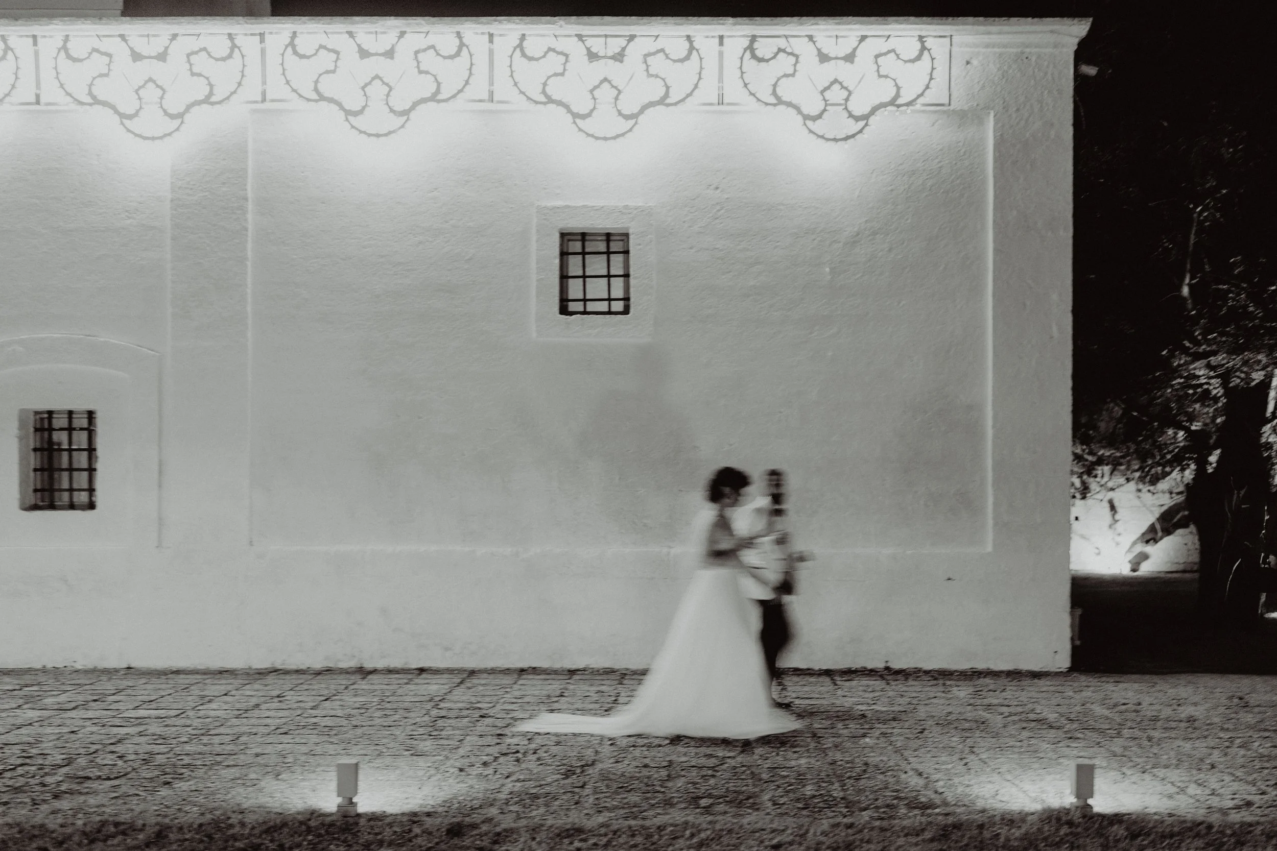 Sposa e sposo che camminano insieme in strada di notte, con sfondo di muro bianco decorato e luci a terra.