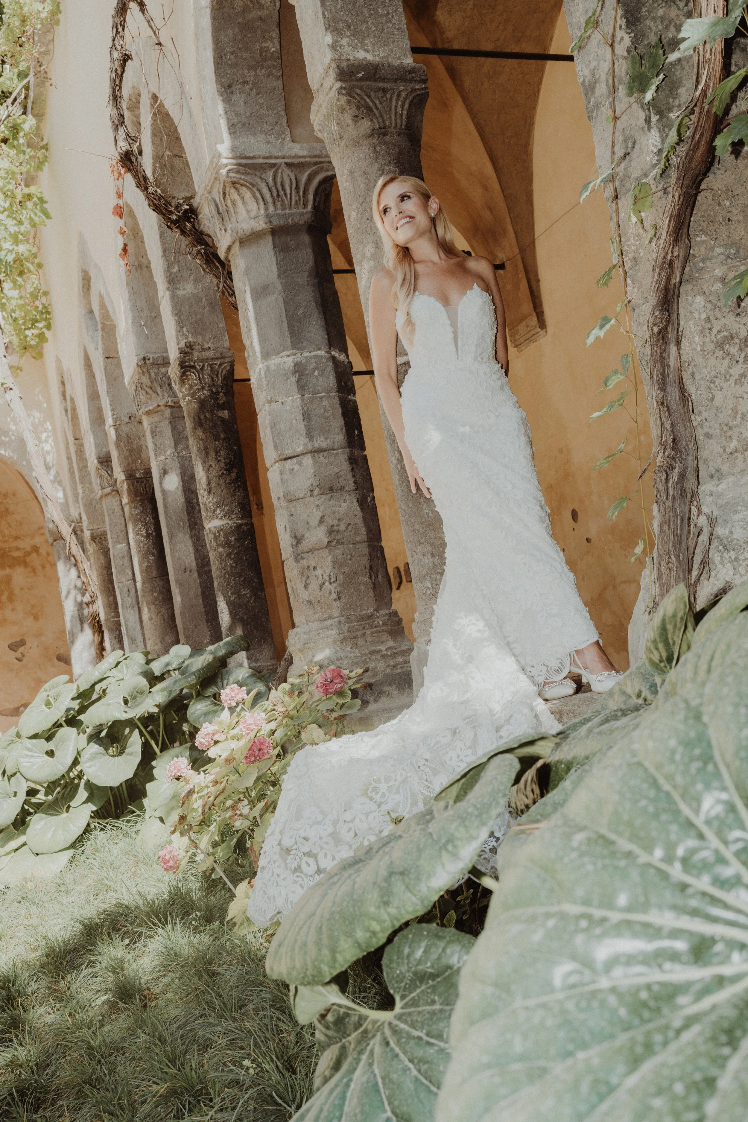 Una sposa indossa un abito da sposa bianco all'interno di un antico edificio con colonne di pietra, circondata da piante verdi e fiori rosa.