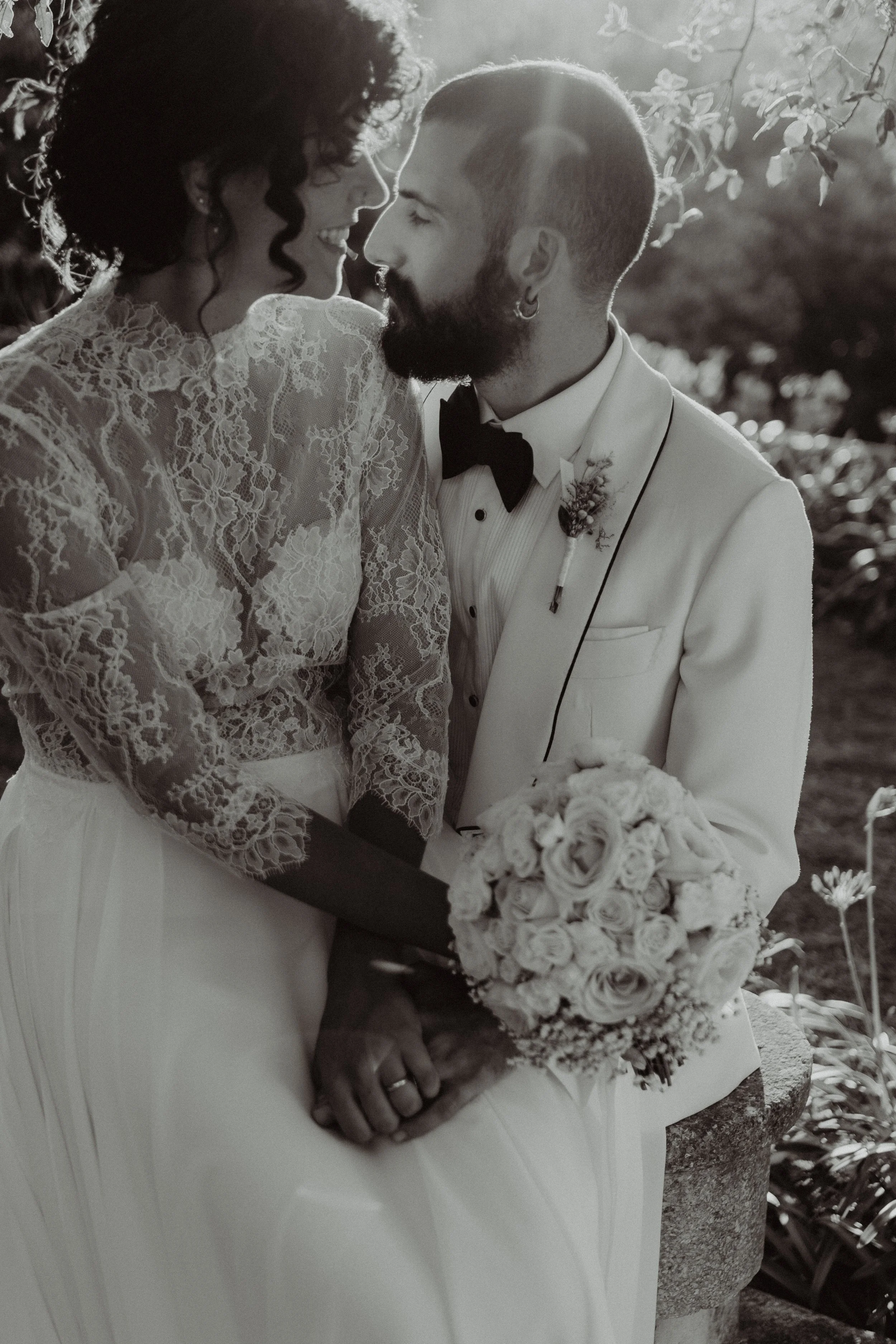 Una coppia felice di sposi si siede su una panchina all'aperto, si tengono per mano e si sorridono, con il volto molto vicino, mentre si scambiano uno sguardo affettuoso, in un contesto naturale. La donna indossa un vestito da sposa con ricami in piz