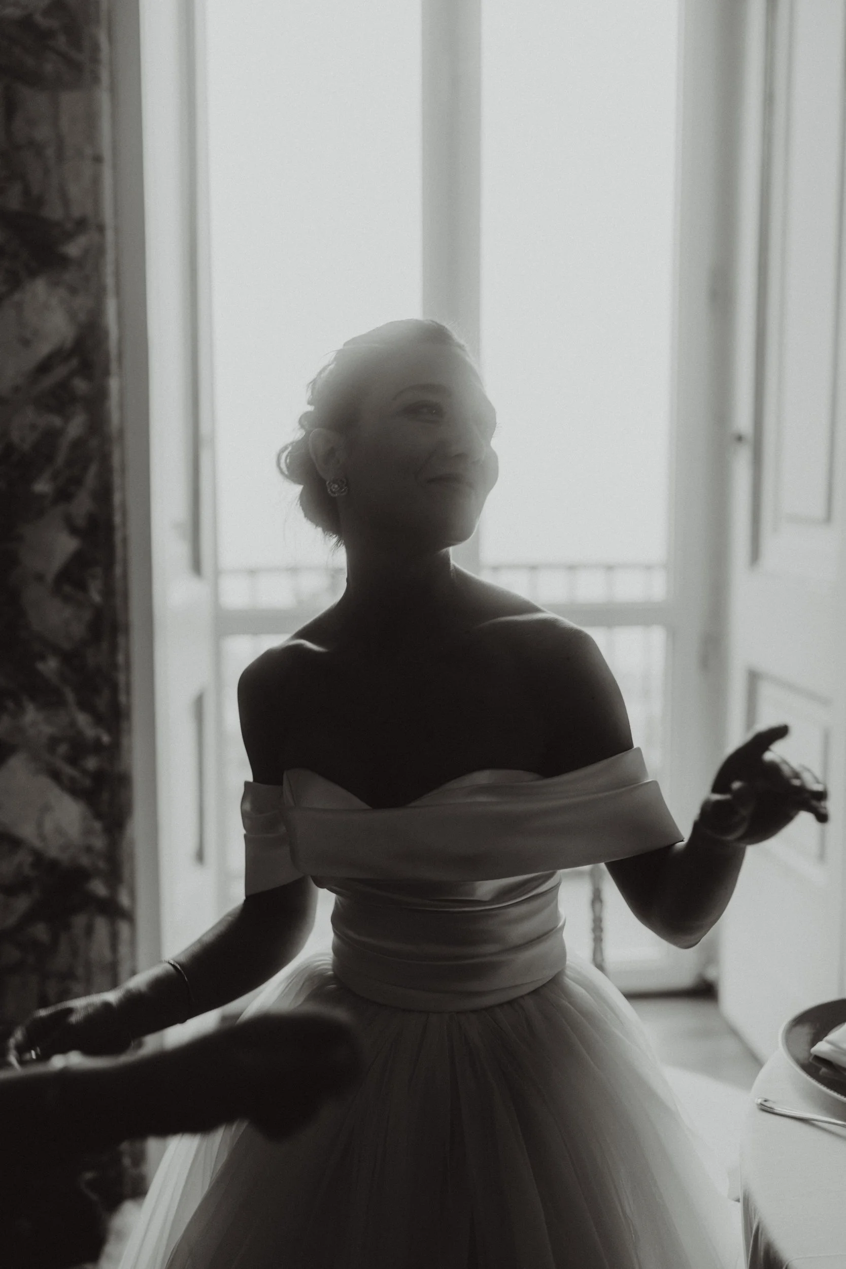 Donna in abito da sposa, vista in controluce di fronte a una finestra luminosa, con una parete di pietra alla sua sinistra.