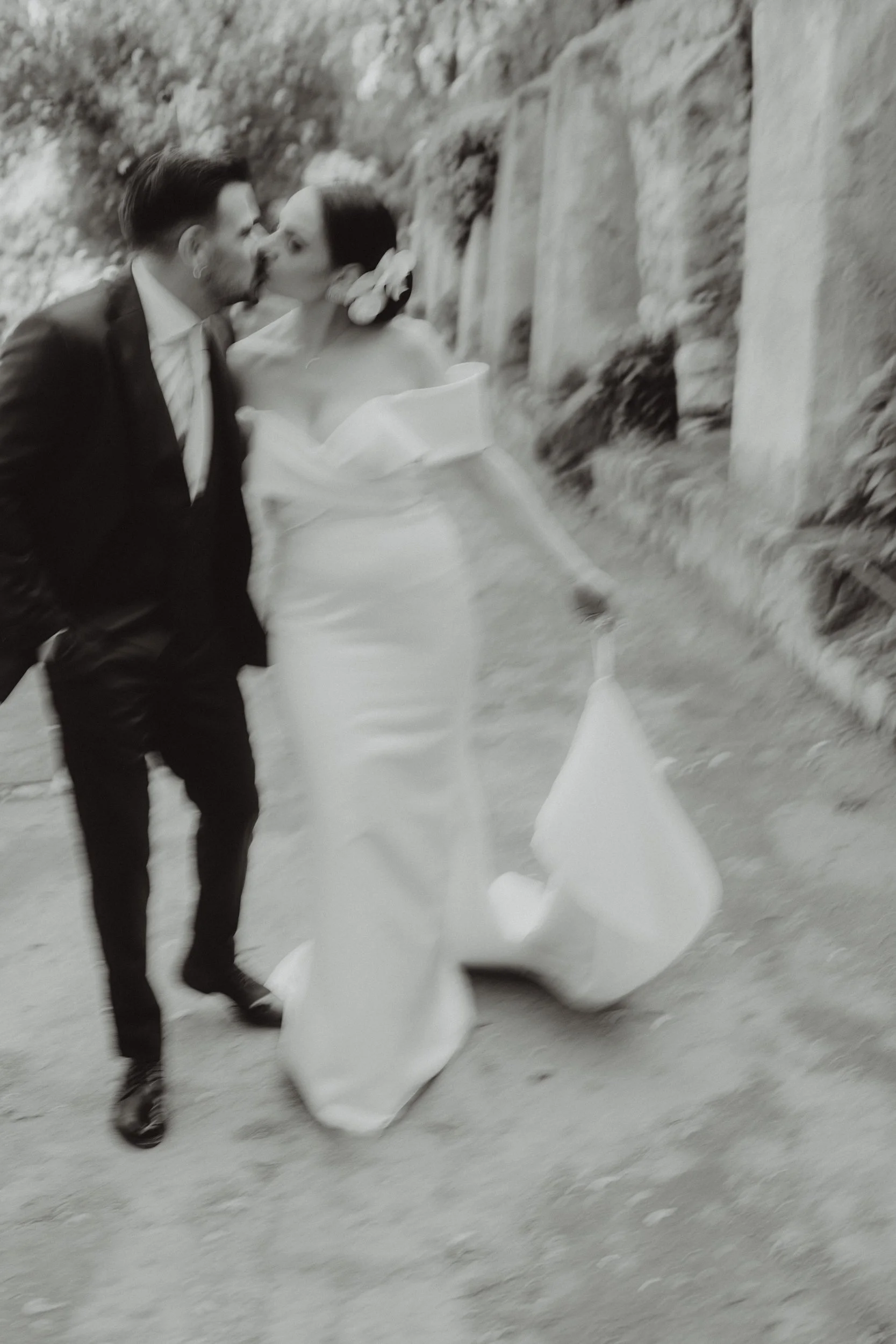 Due persone, un uomo in abito e una donna in abito da sposa, si baciano con uno sfondo naturale, probabilmente un sentiero di montagna o di campagna.