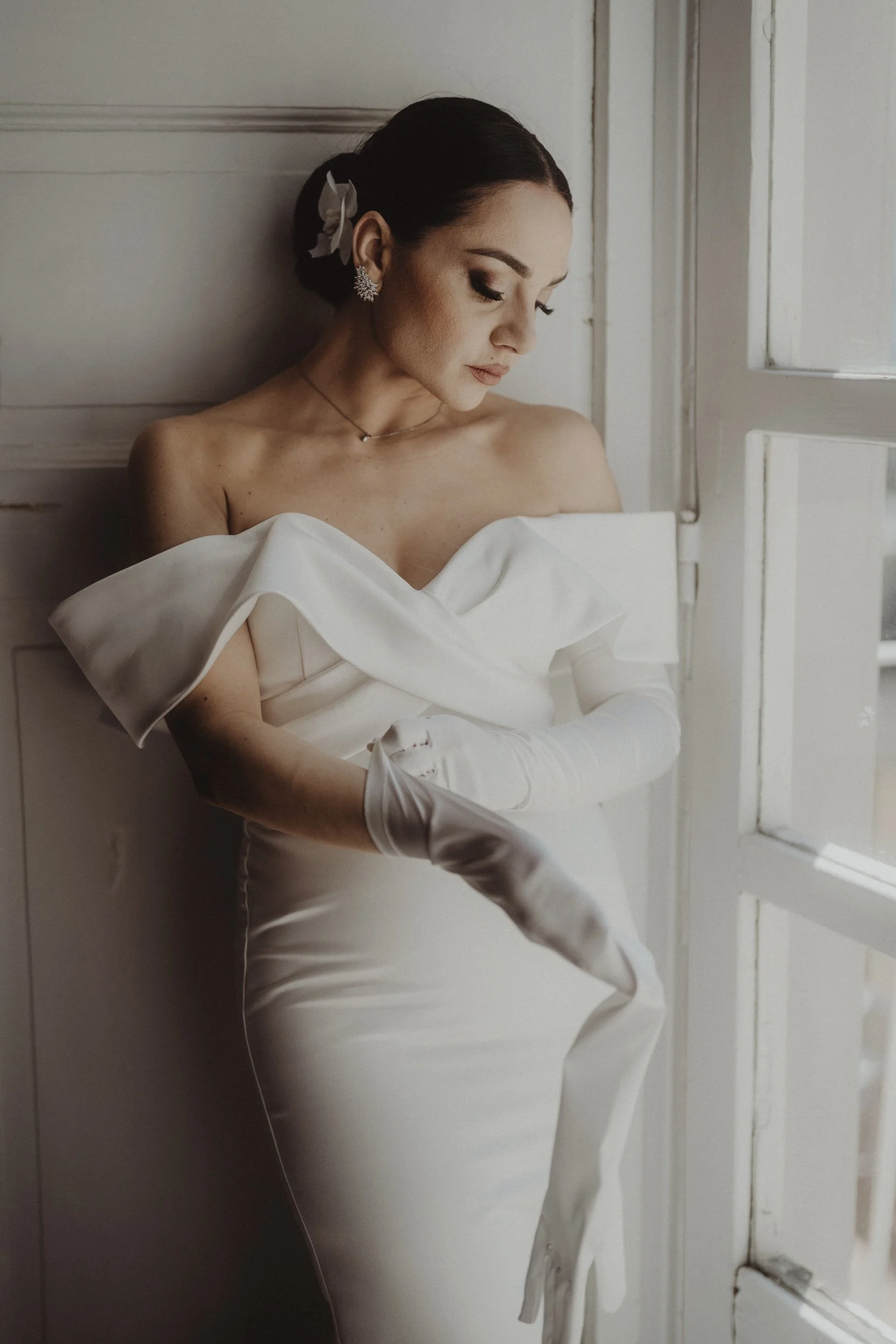 Vechia donna elegante con vestito bianco e guanti lunghi, posa vicino a una finestra in una stanza con pareti chiare, capelli raccolti con fiocco e orecchini a forma di fiore.