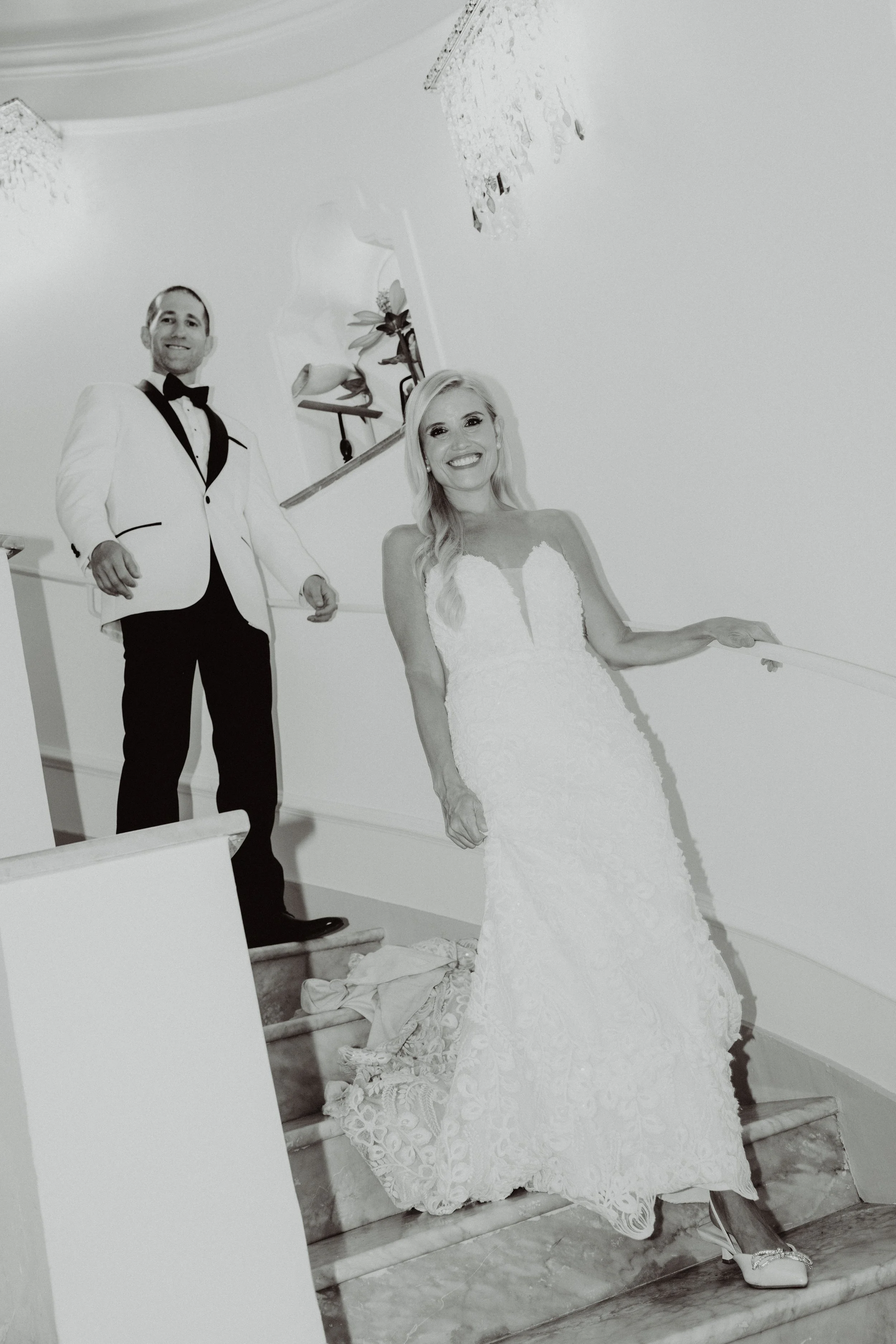 Una coppia di sposi in abiti eleganti si trova su una scala di marmo in un ambiente interno, con decorazioni e lampadari, sorridenti durante il loro matrimonio.
