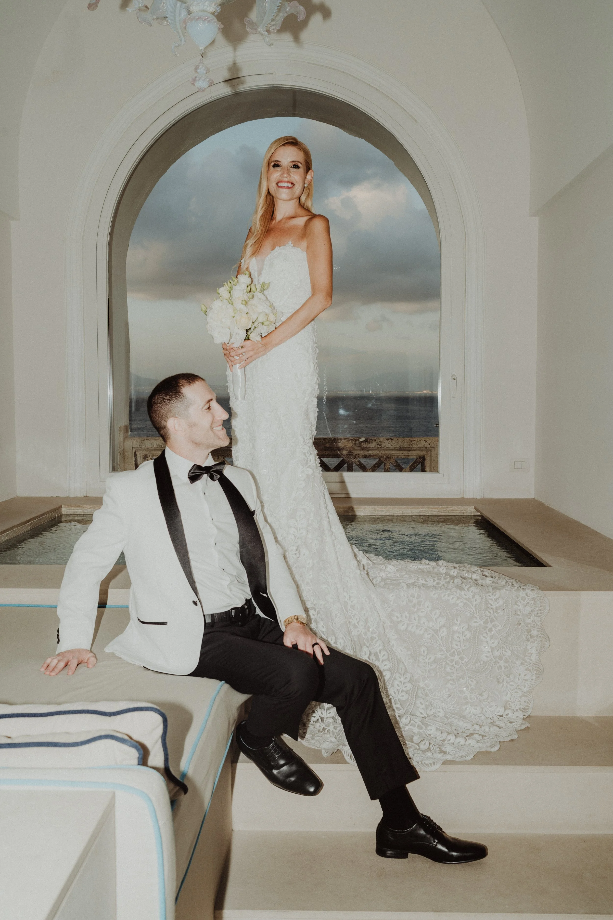 Una coppia di sposi, con la donna in abito da matrimonio e l'uomo in smoking, posa in una sala con vista sul mare, durante il tramonto.