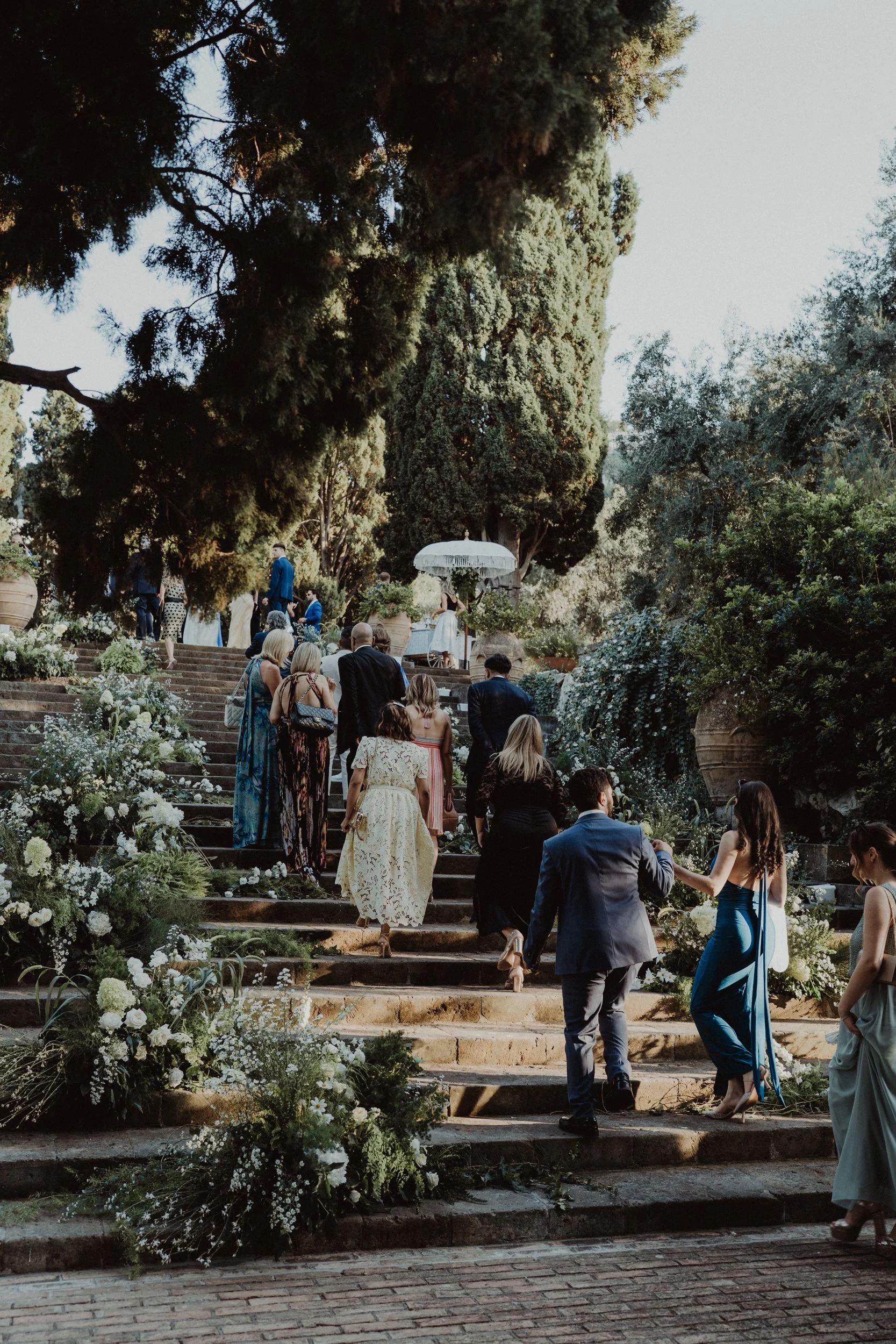 Persone che salgono le scale decorati con fiori in un giardino all'aperto, probabilmente durante un matrimonio o un evento formale, con alberi e vegetazione sullo sfondo.