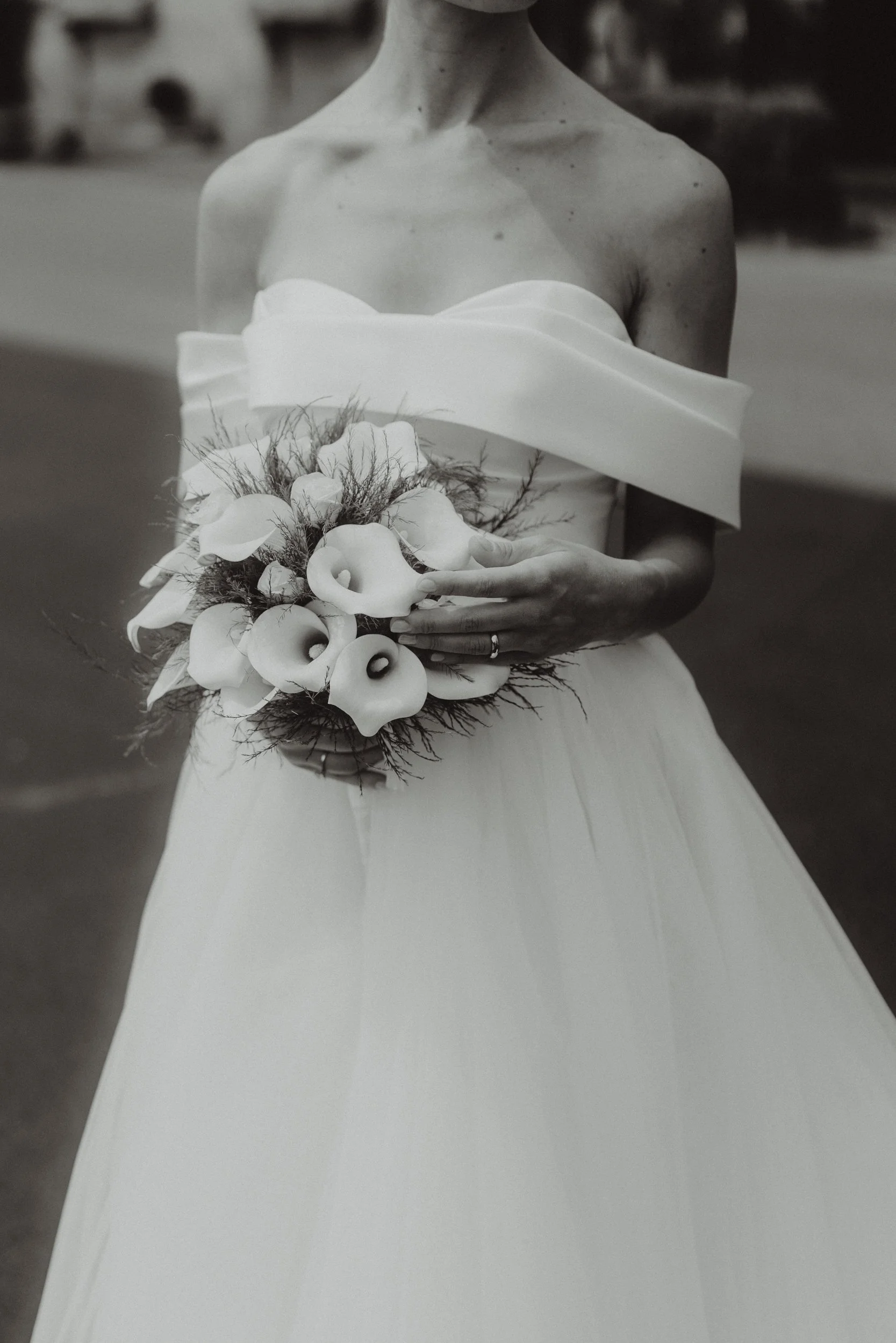 Una donna in abito da sposa bianco tiene in mano un bouquet di fiori bianchi e calla, con sfondo sfocato.