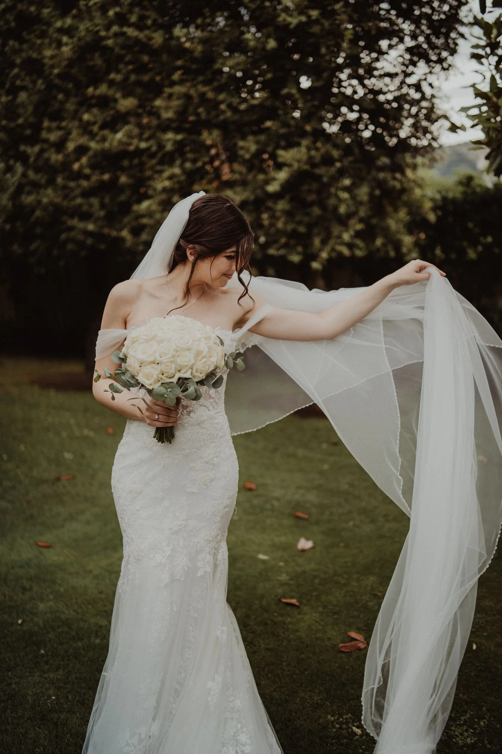 Una sposa in abito da sposa bianco con foulard e velo, che tiene un bouquet di rose e foglie, in un giardino con alberi e erba verde, in un'ambientazione naturale.