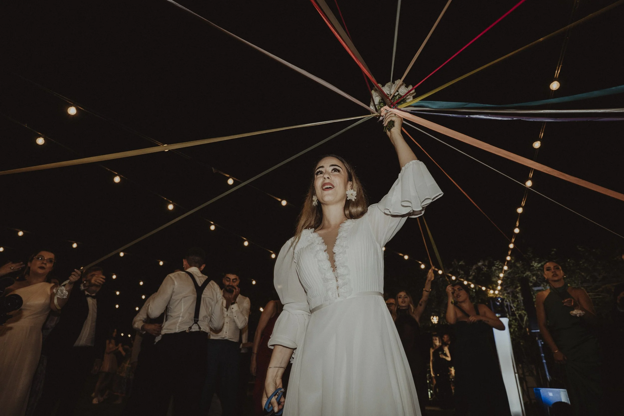 Una donna in abito da sposa tiene una lampada decorativa colorata durante una festa serale, circondata da altre persone sotto luci appese.