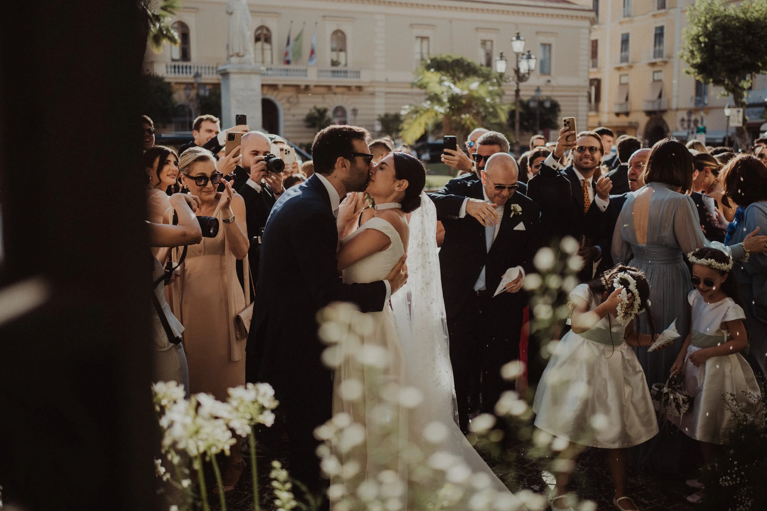 Una coppia di sposi si baciano circondata da amici e parenti in un matrimonio all'aperto, con alcuni bambini vestiti da damigelle.