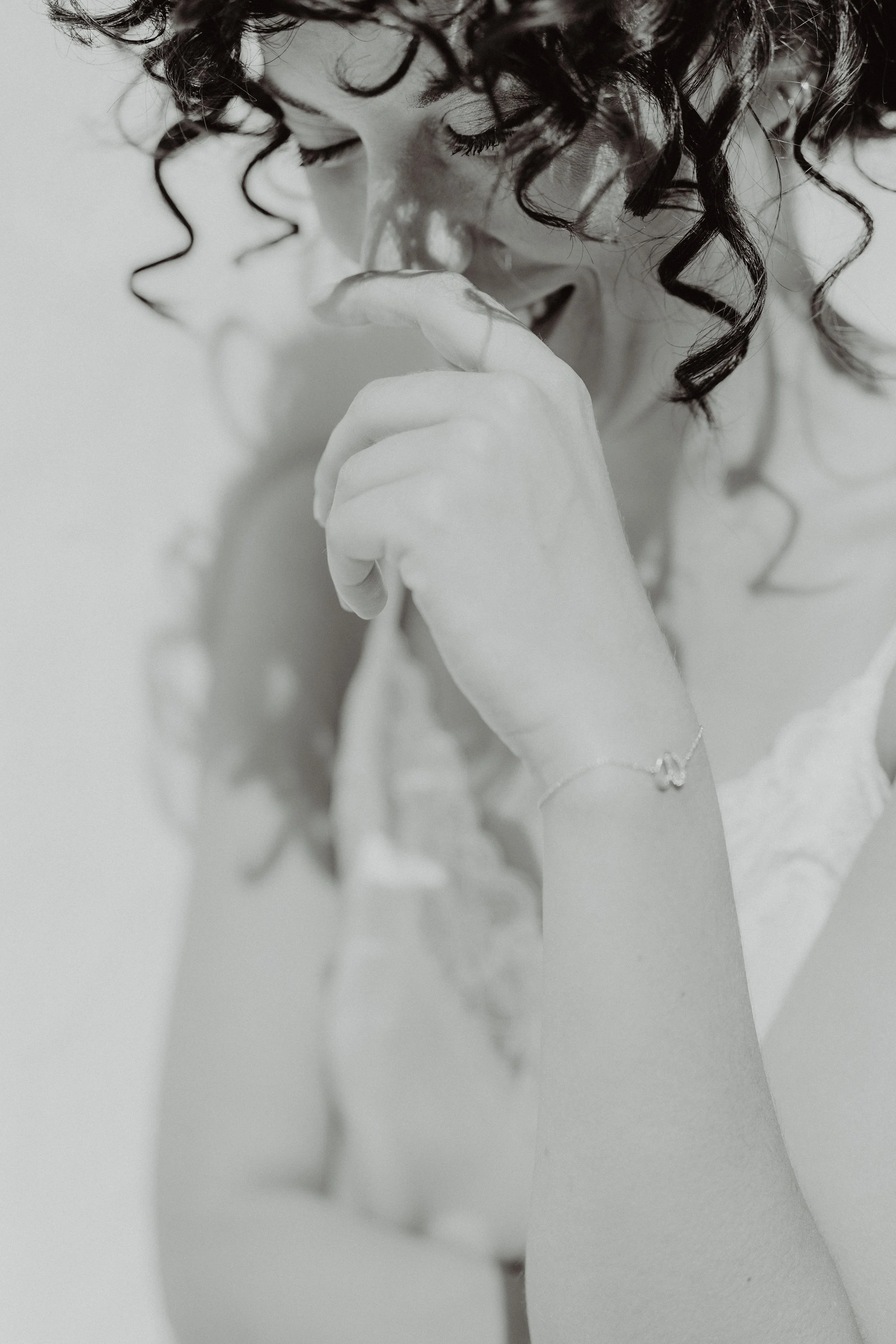 Ragazza con capelli ricci, sorriso, mano vicino al viso, indossa braccialetto e vestito bianco.