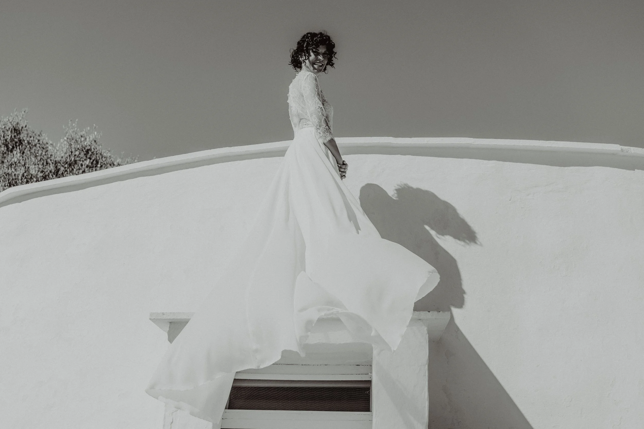 Una donna in abito da sposa si trova su un balcone bianco, con il sole che proietta la sua ombra sulla parete.