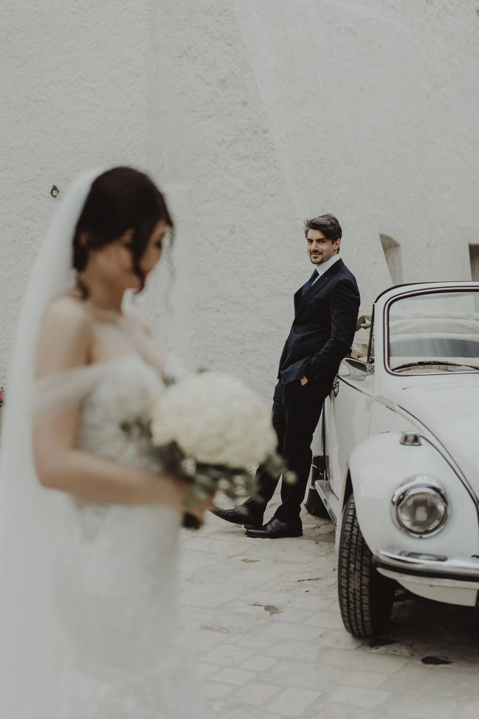 Sposo che guarda la sposa con bouquet, in primo piano la sposa con abito da matrimonio e velo, sullo sfondo un'auto d'epoca bianca