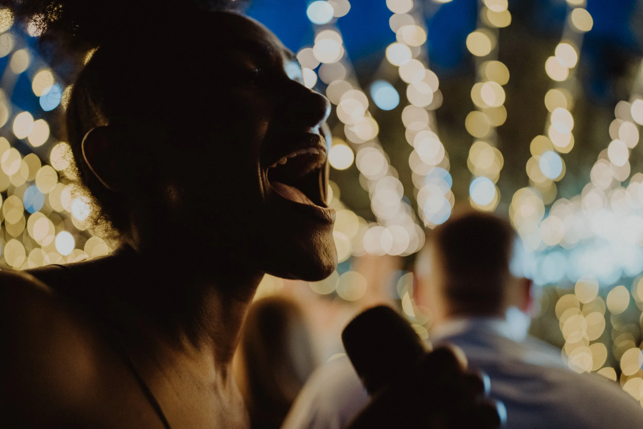 Una donna afroamericana che canta o parla con gioia, tenendo un microfono, con luci di feste sfocate sullo sfondo.