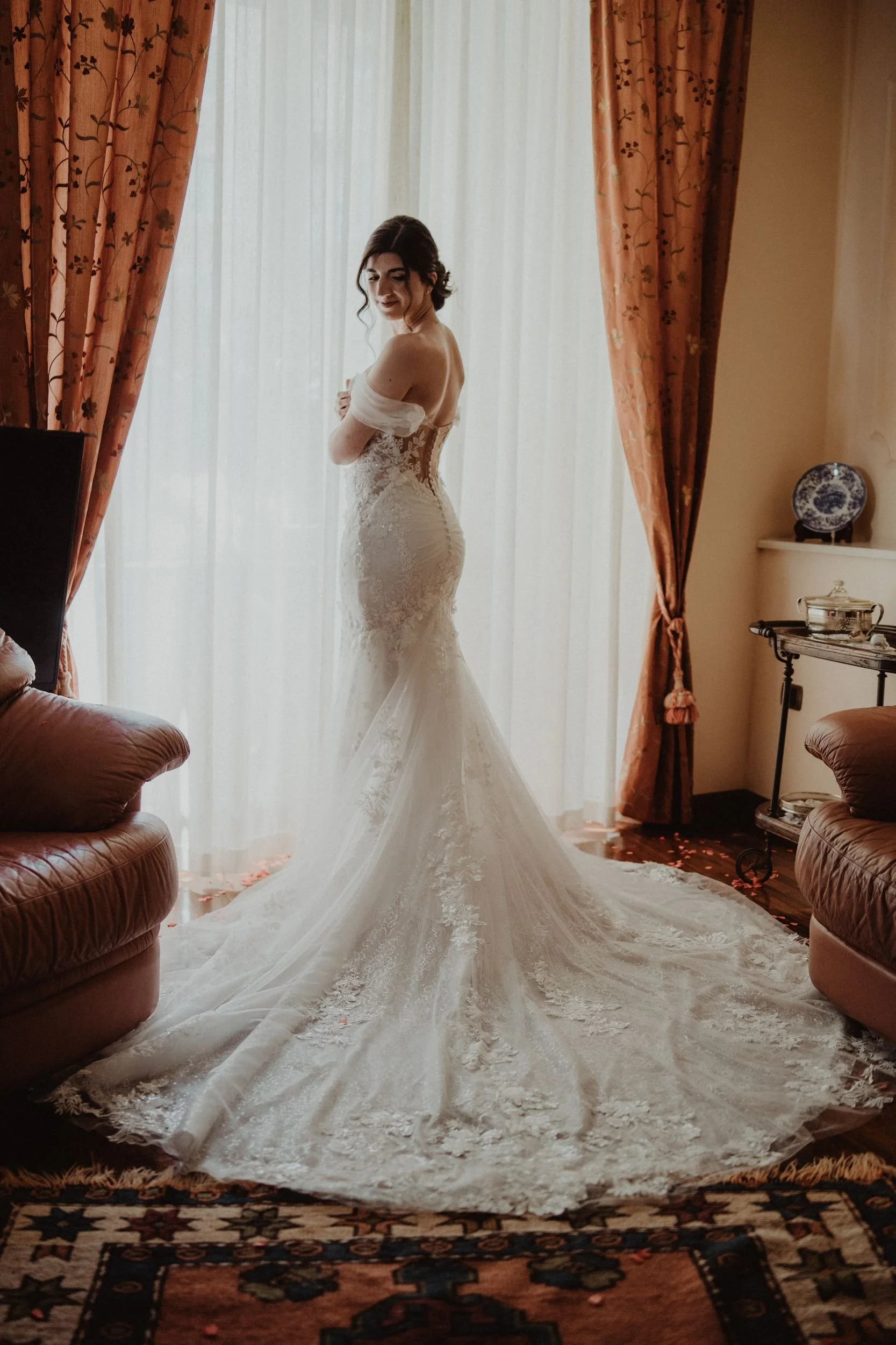 Una sposa in abito da nozze bianco con dettagli in pizzo si trova in una stanza illuminata dalla luce naturale che entra da una finestra con tende di colore rosa antico. La sposa si sta sistemando mentre guarda leggermente di lato, con un'espressione